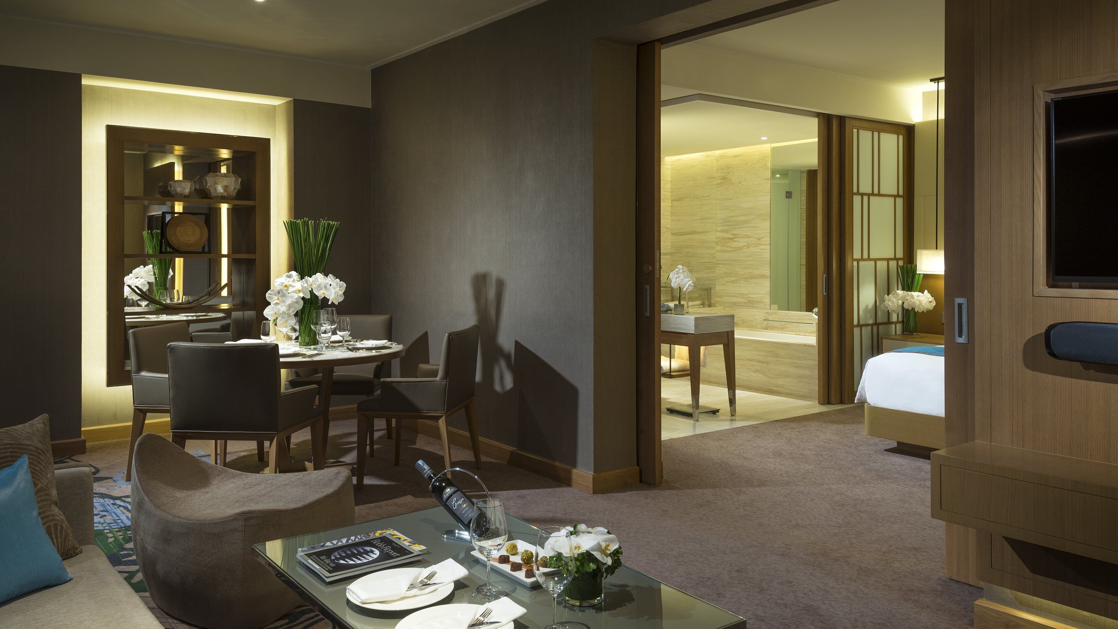 A hotel room with a dining area, a sitting area with a sofa and coffee table, and a bedroom with a bed and a TV.