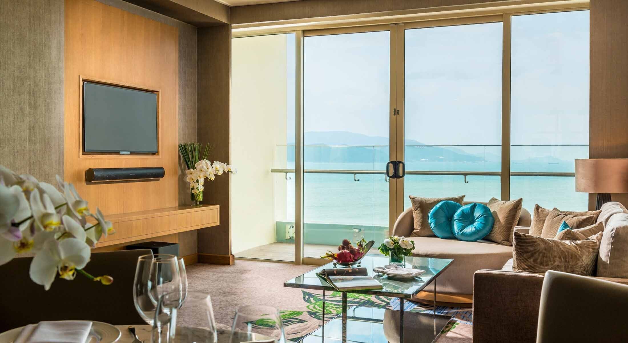 A hotel room with a large window, a flat-screen TV, a sofa with cushions, a glass coffee table, and a dining area with a table set for two.