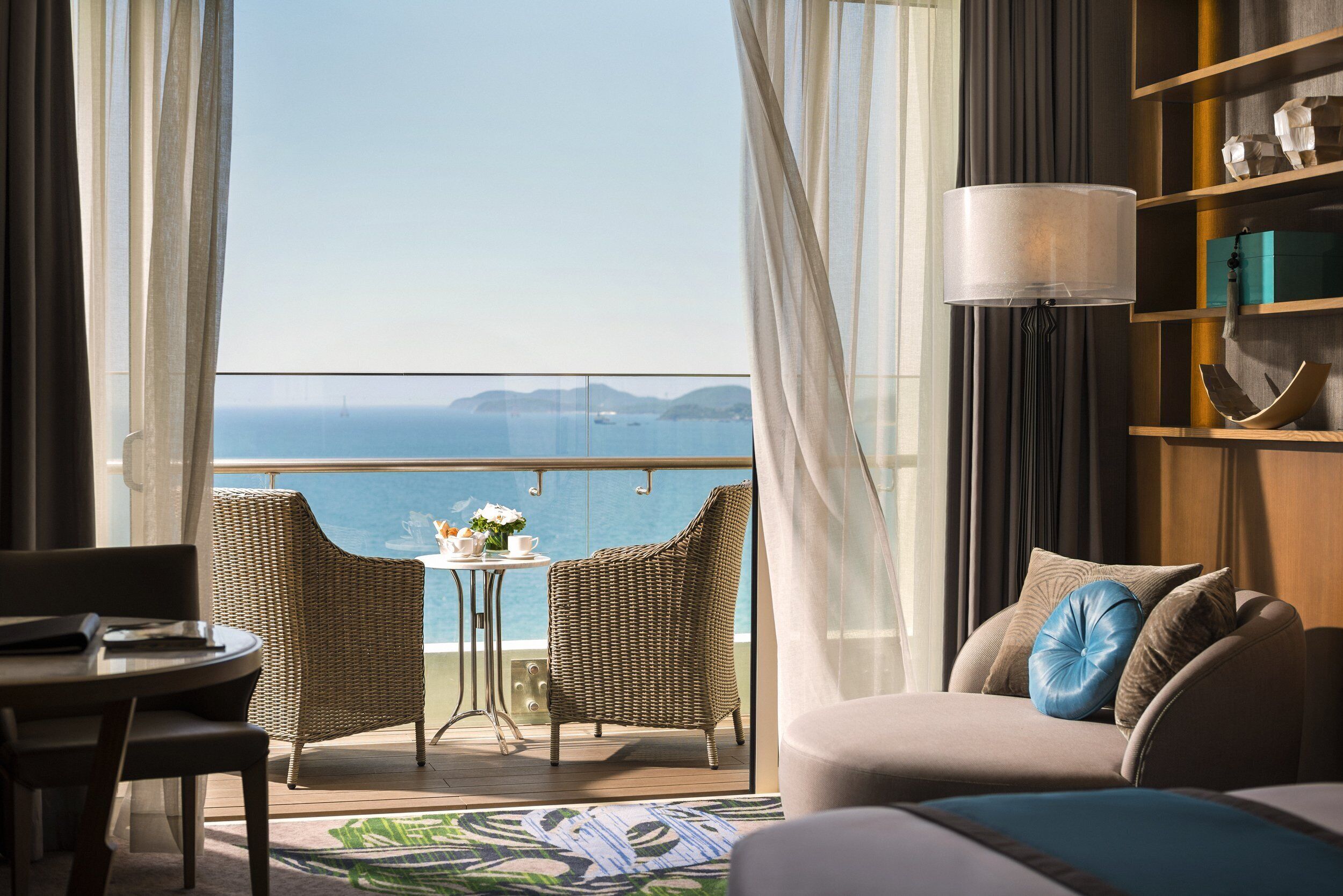 Un balcon avec des fauteuils en osier, une table ornée d’un vase et une vue sur la mer.
