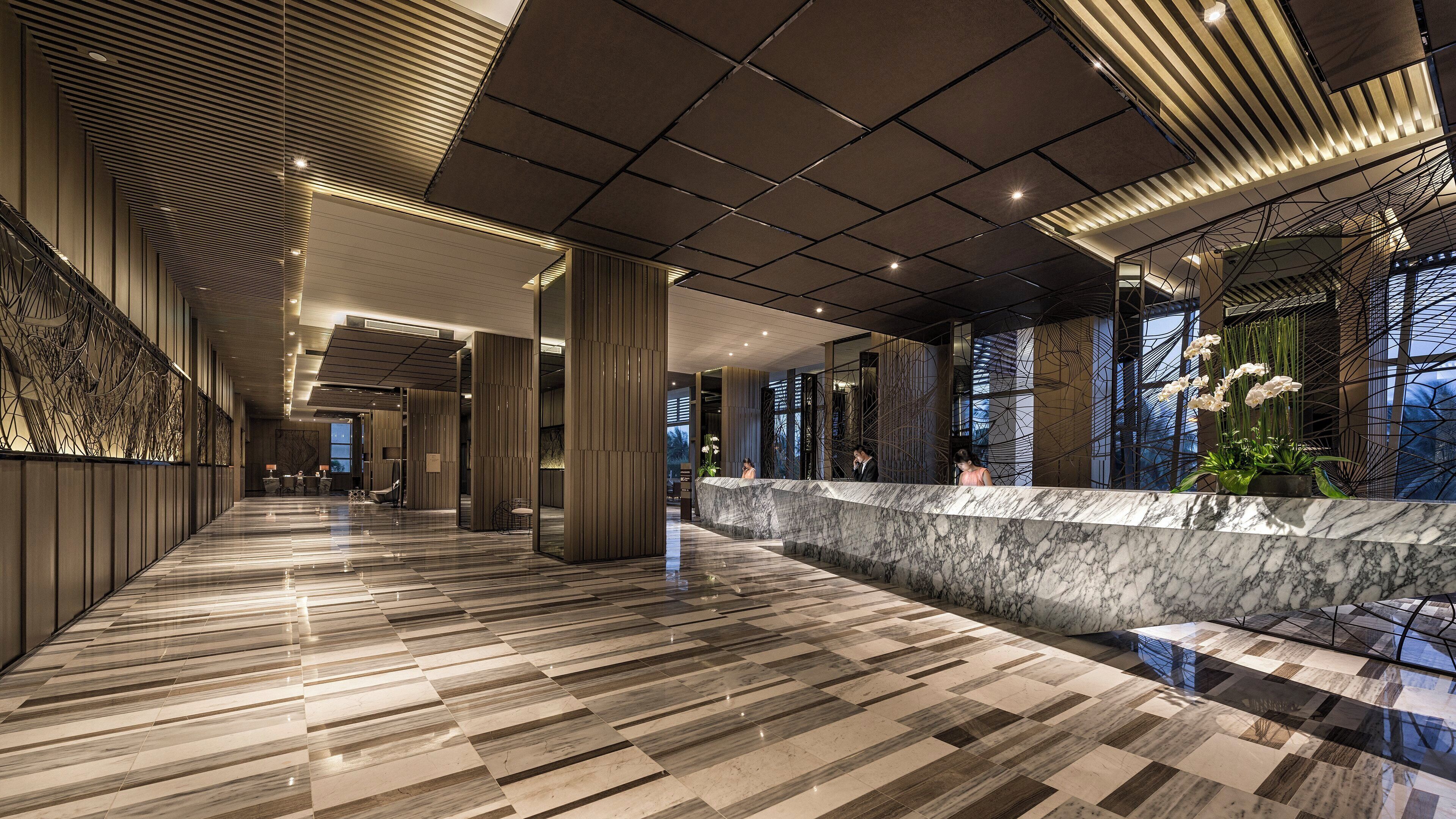 Un hall d’hôtel moderne avec un comptoir d’accueil en marbre, un long couloir au sol décoré de motifs et un plafond orné de motifs géométriques.