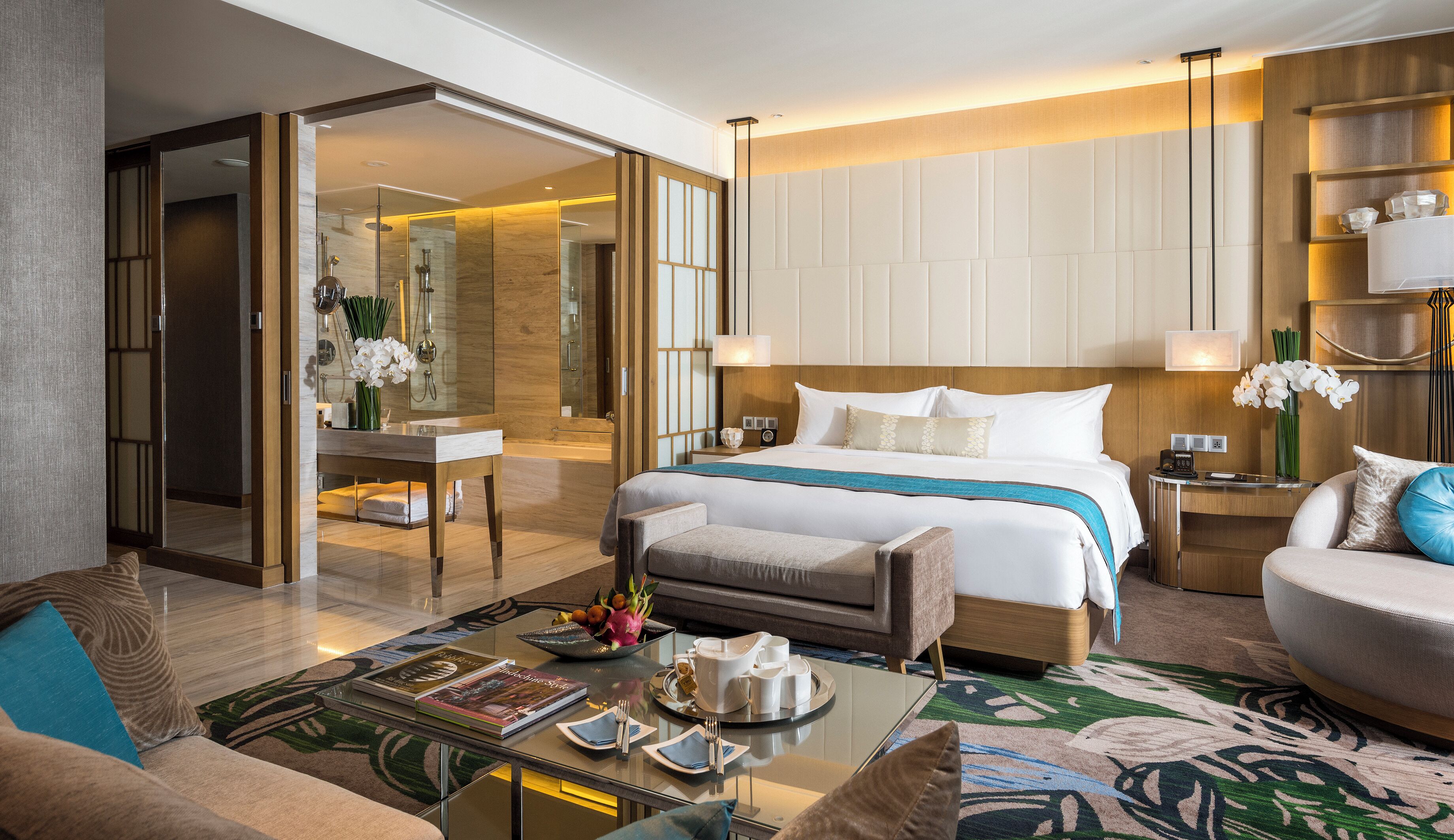 A modern hotel room with a large bed, a sitting area with a sofa and armchair, a glass coffee table, and a dining area with a tray of breakfast items.