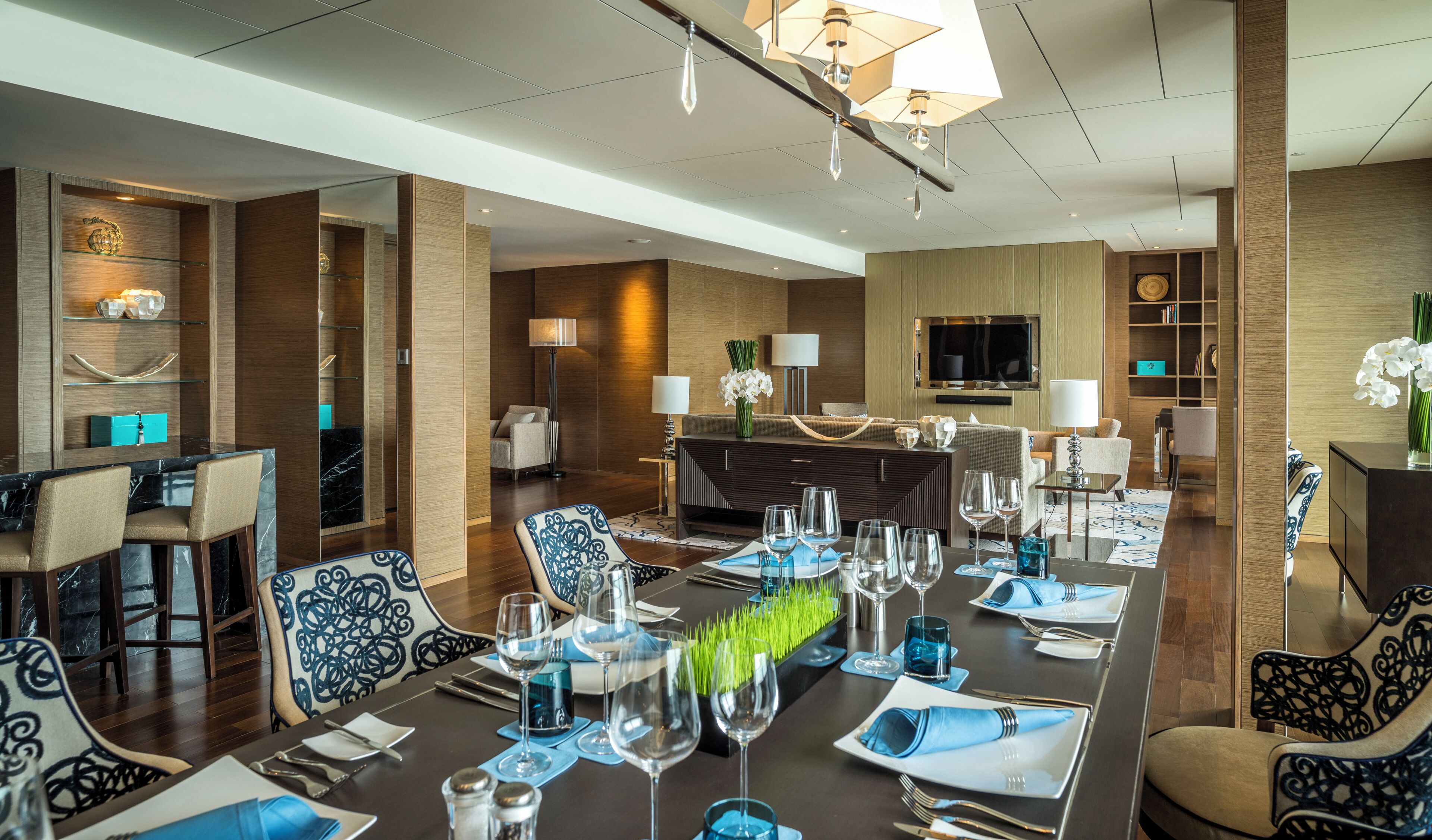 A modern hotel dining area with a long table set for a meal, a bar area, and a seating area with sofas and a TV.