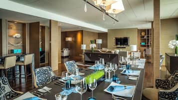 A modern hotel dining area with a long table set for a meal, a bar area, and a seating area with sofas and a TV.