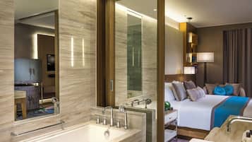 A hotel room with a large mirror, a bathtub, a bed with multiple pillows, and a bedside table.