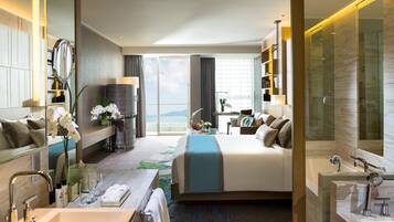 Een moderne hotelkamer met een groot bed, een zithoek met een bureau, een badkamer met bad en douche, en uitzicht op de zee door het raam.