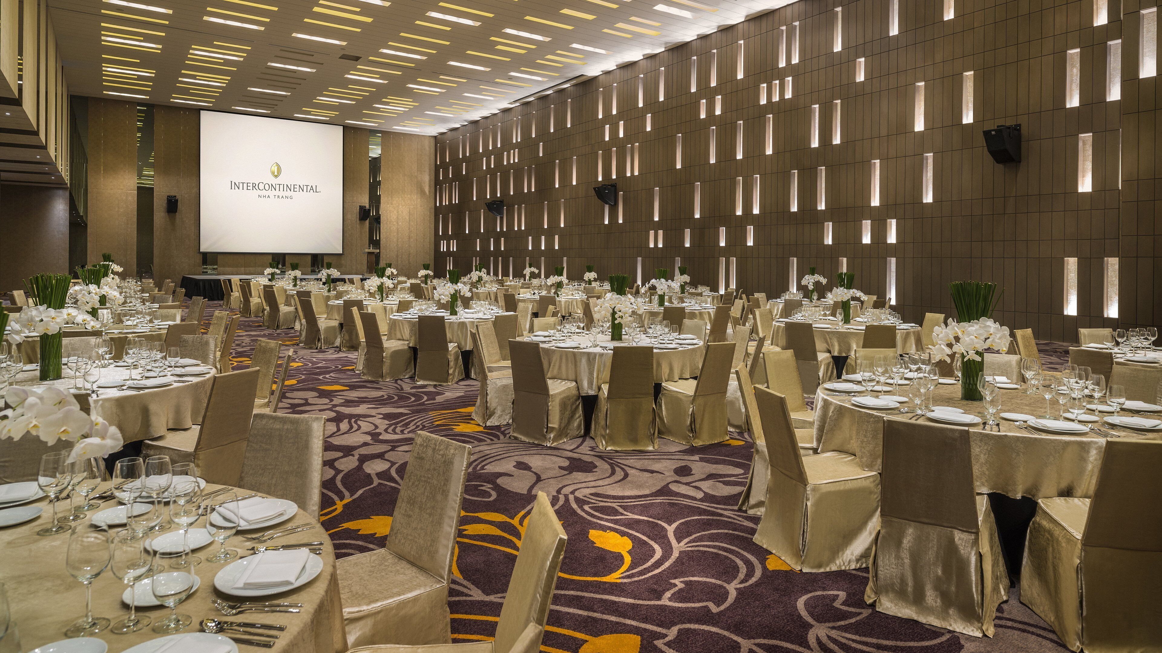 A banquet hall with round tables set for a formal event, featuring a carpeted floor and a backdrop with a projection screen.