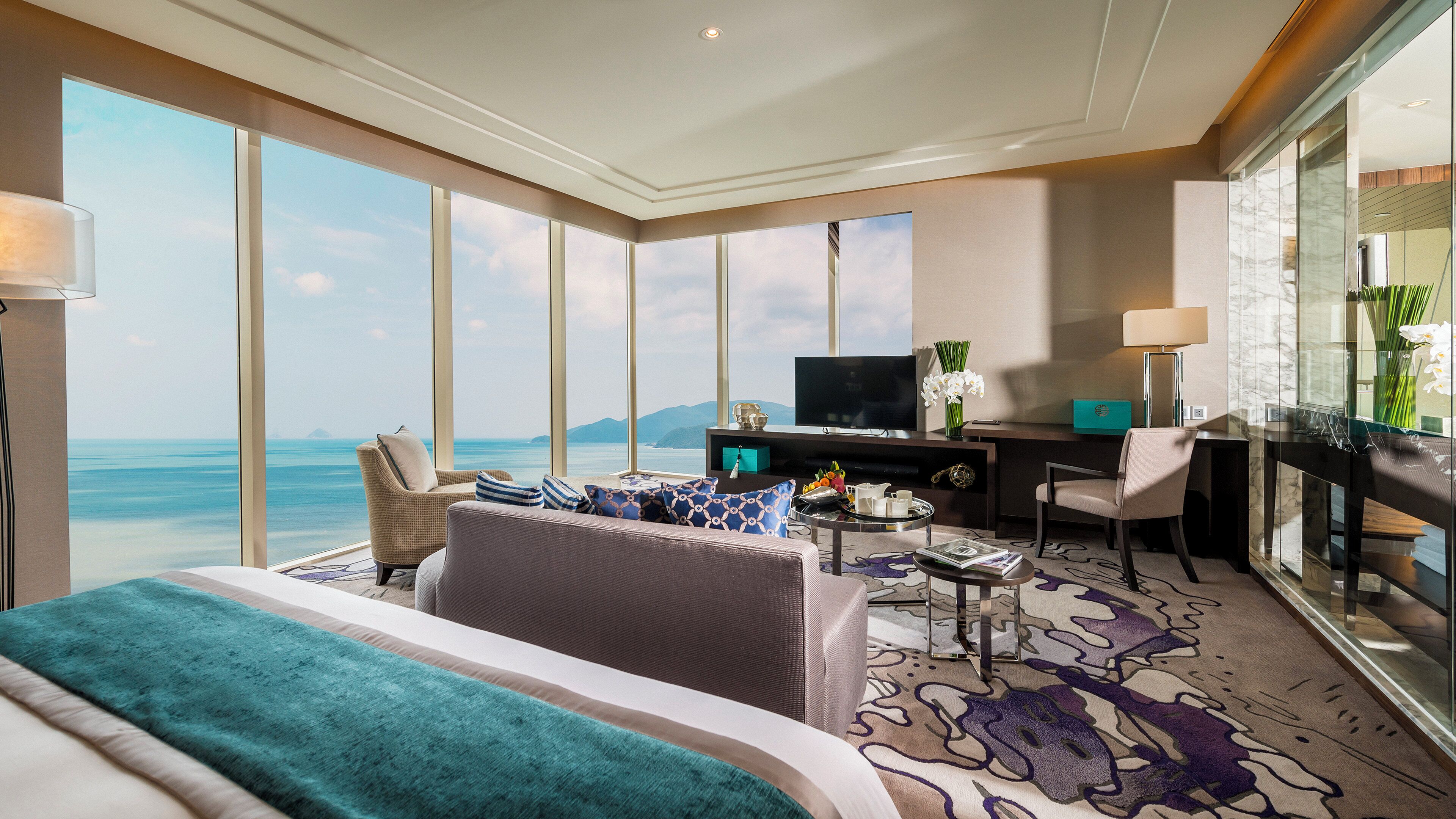 A hotel room with a large bed, a desk with a TV, and a view of the sea.