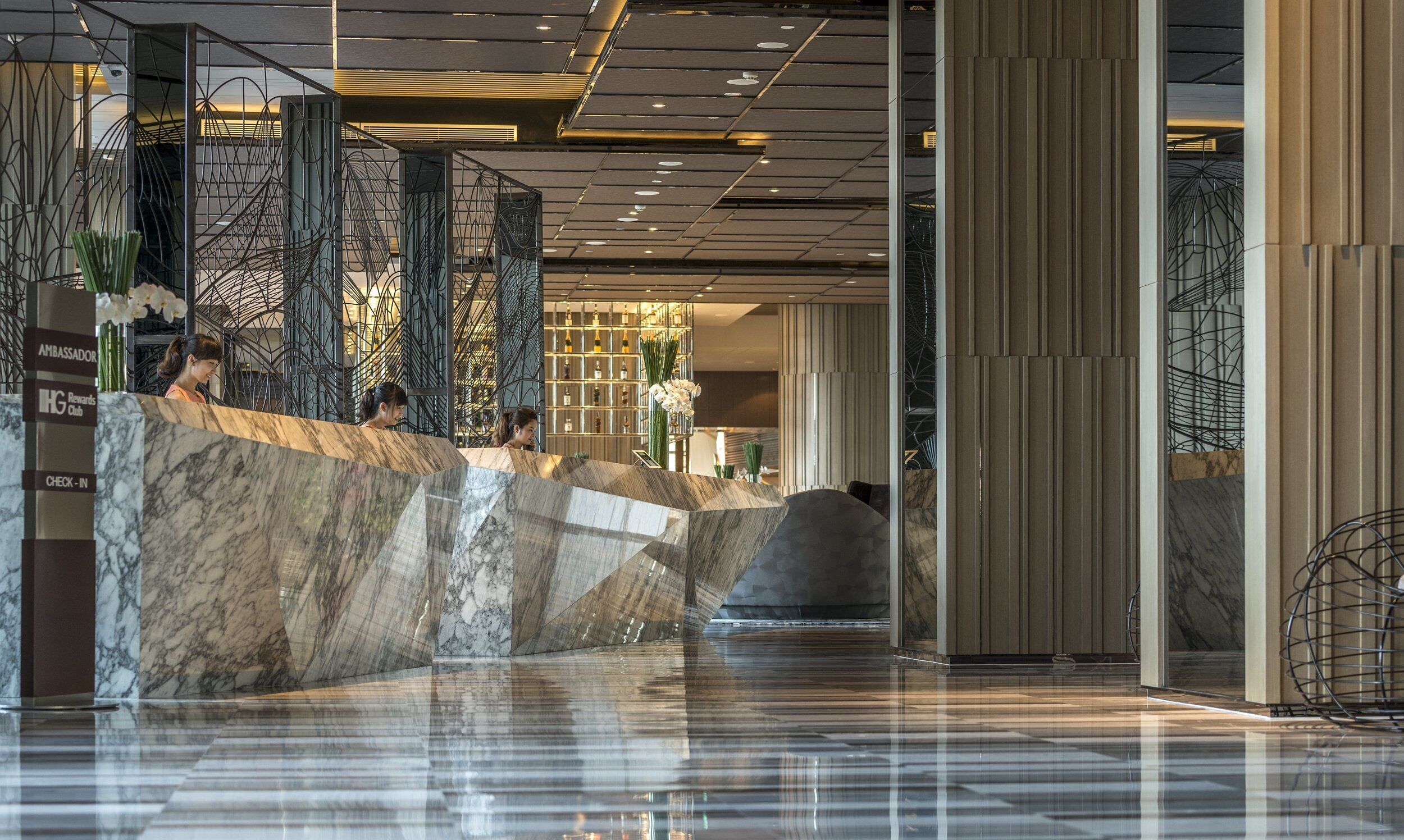 A modern hotel lobby with marble reception counters, a check-in area, and a bar with a wine display.