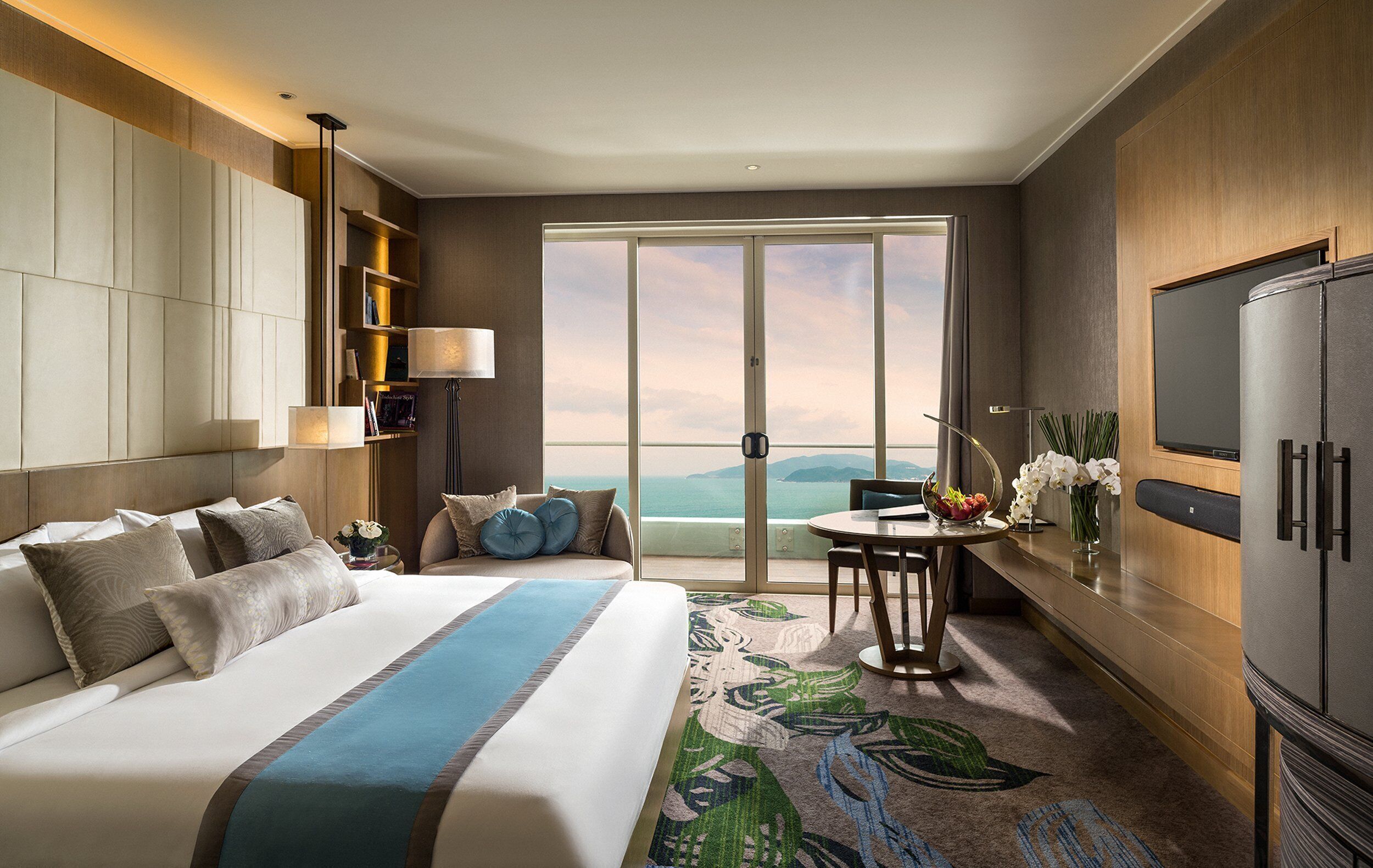 A hotel room with a large bed, a desk with a chair, a television, and a view of the ocean.