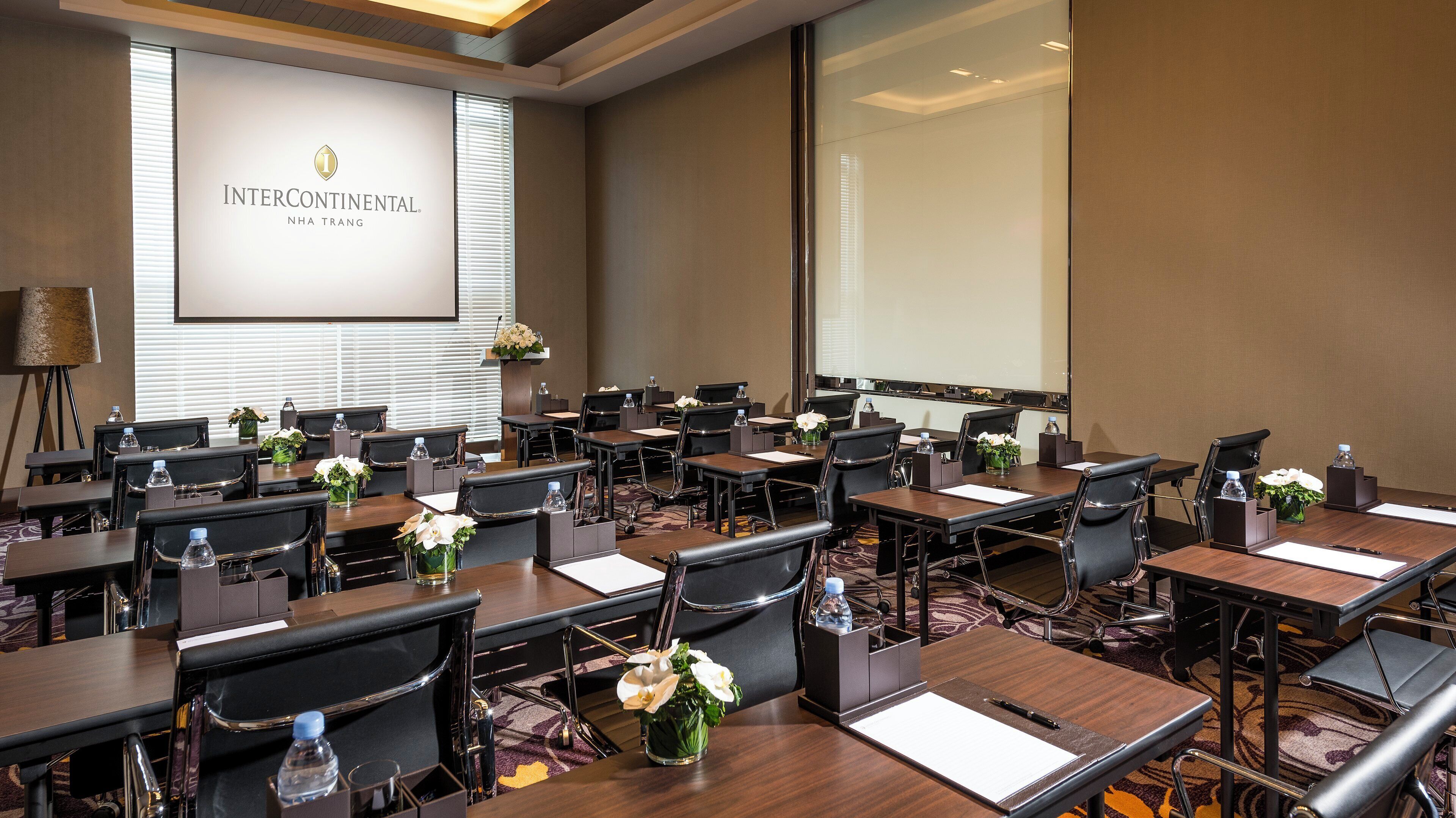 A conference room with multiple tables, chairs, and a large screen displaying the hotel name.