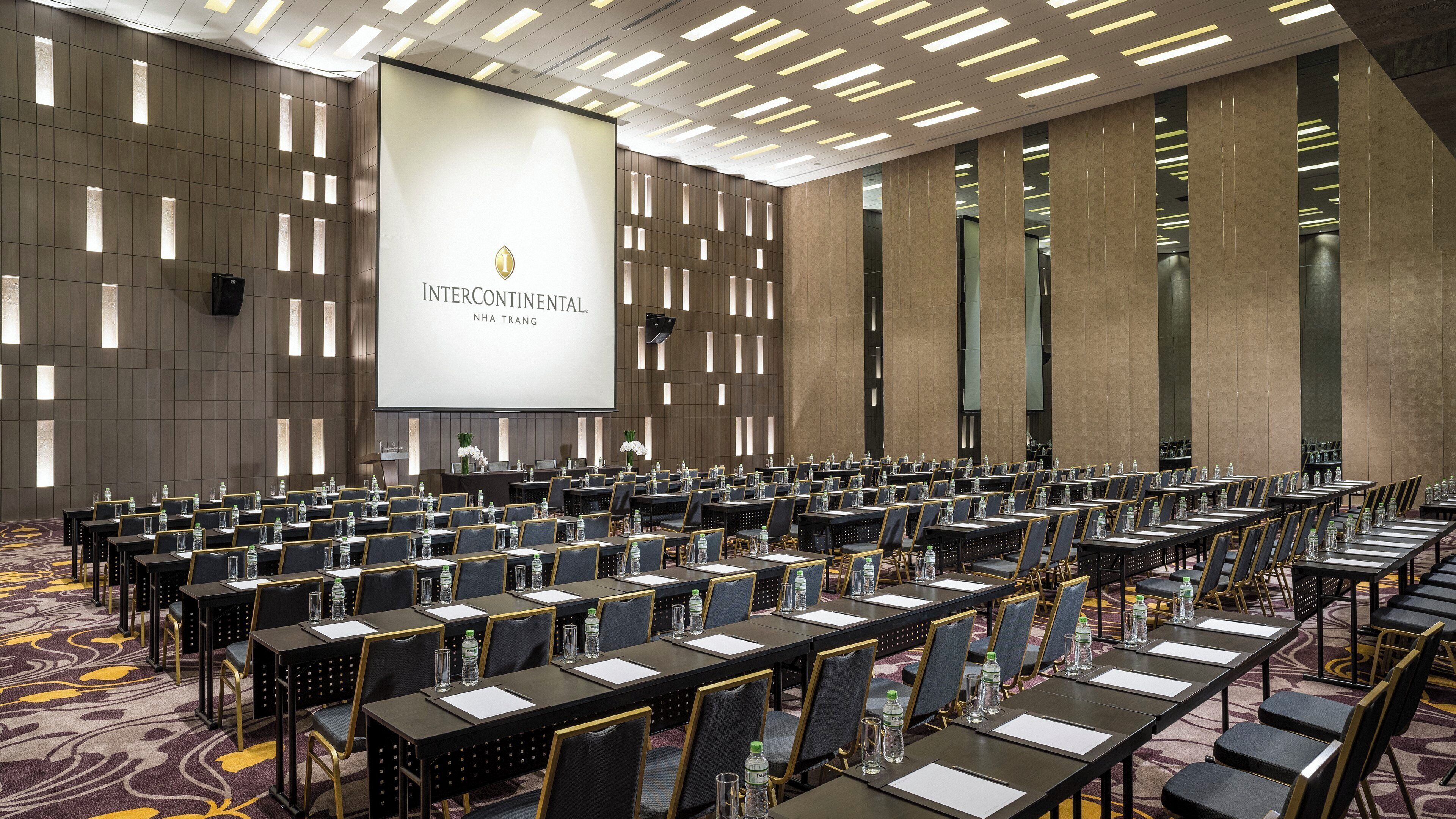 A conference room with rows of tables and chairs, a large screen displaying 'INTERCONTINENTAL NHA TRANG', and a modern ceiling design.