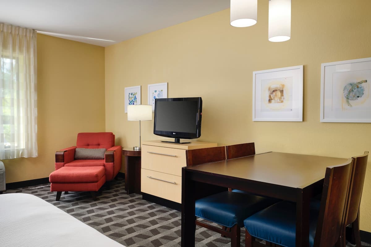 Une chambre d’hôtel avec un lit, un fauteuil rouge, une table à manger en bois avec des chaises bleues, un téléviseur posé sur un meuble et deux tableaux encadrés accrochés au mur.