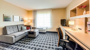 Ein Hotelzimmer mit Sofa, Schreibtisch, Stuhl, Fernseher, Lampe und einem Fenster mit Vorhängen.