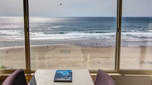 A room with a large window overlooking a beach. There is a table with a book on it and two chairs. - Seahorse Oceanfront Lodge (Lincoln City)