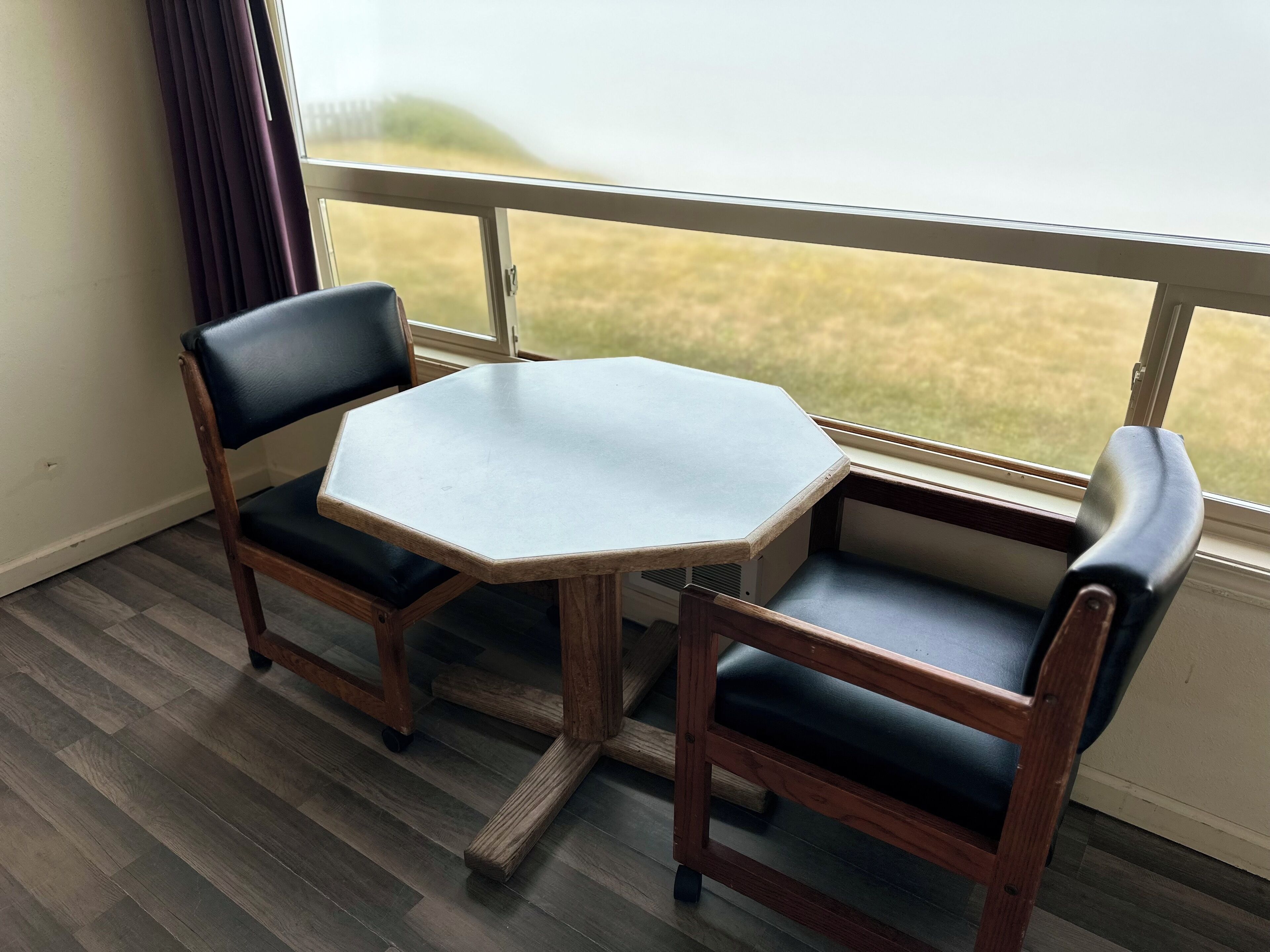 A small octagonal table with two chairs, a window with a view of a grassy field, and wooden flooring.