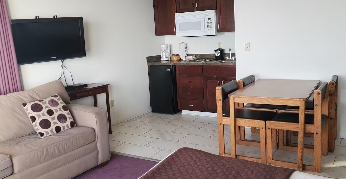A compact living space with a sofa, a dining table with chairs, a kitchenette with a microwave, and a flat-screen TV mounted on the wall.