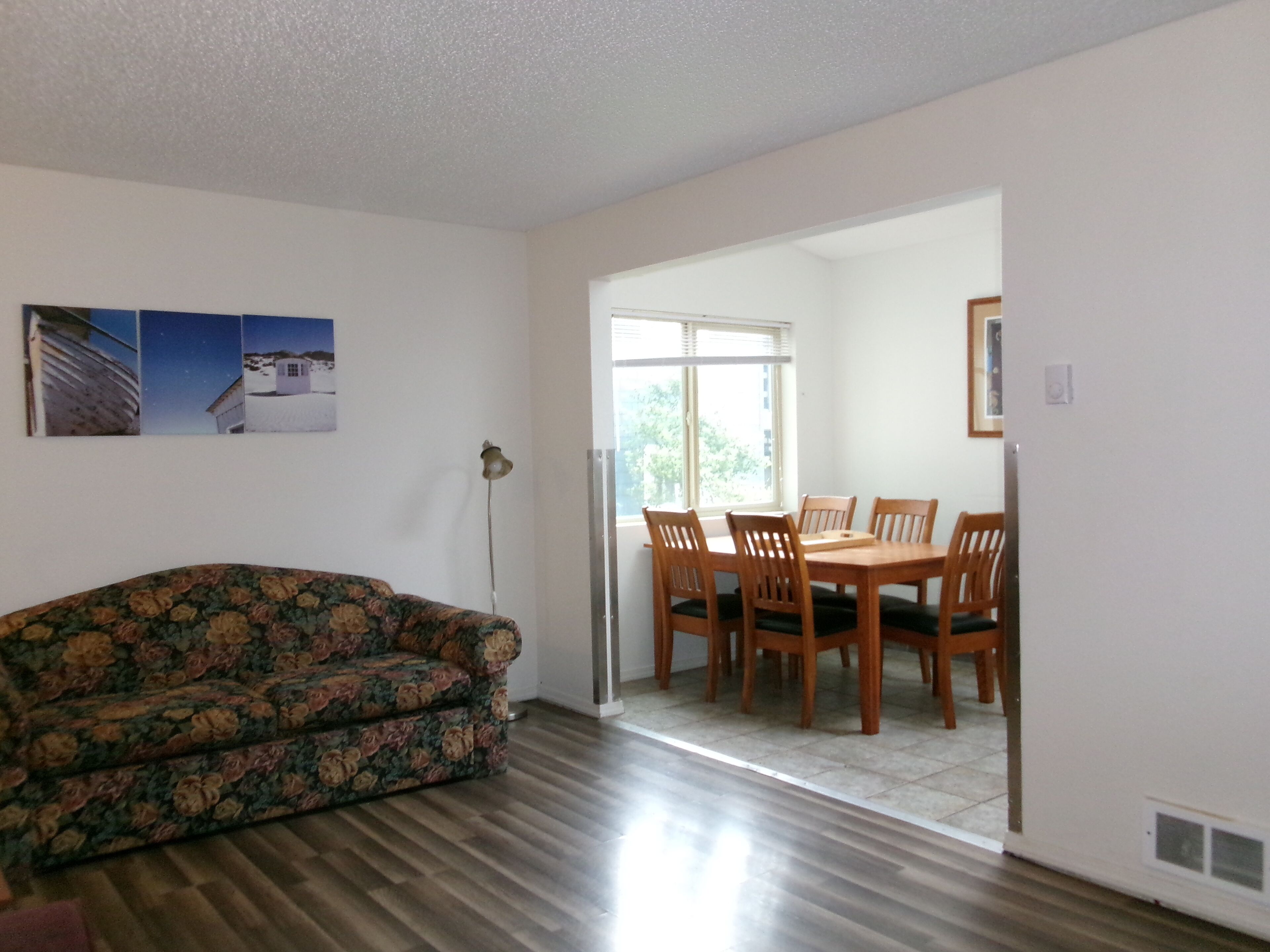 A living room with a floral sofa, a dining table with chairs, and a large window.