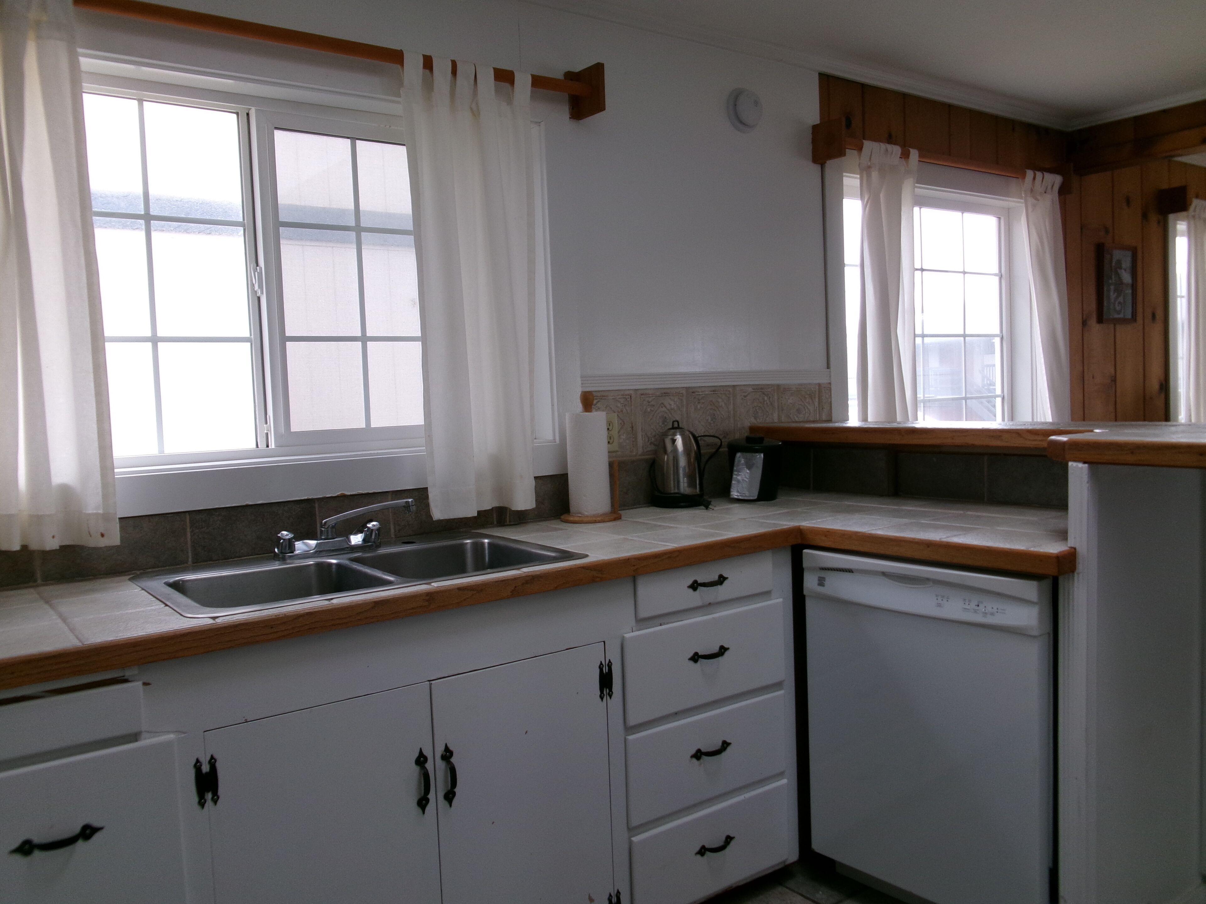 A kitchen with white cabinets, a double sink, wooden countertops, and two windows with white curtains.