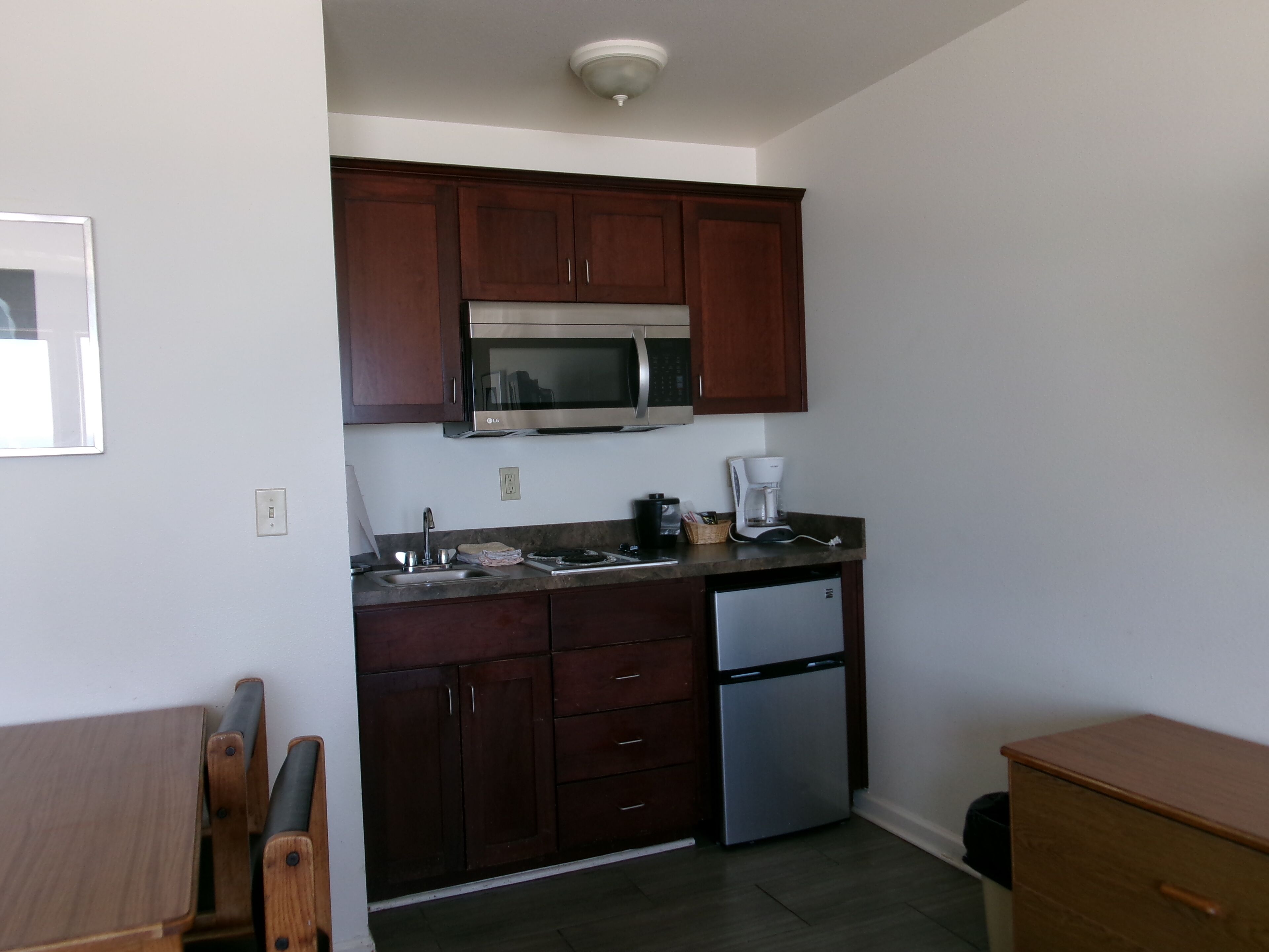 Una cocina compacta con armarios de madera oscura, un microondas de acero inoxidable, horno empotrado, cafetera y refrigerador.