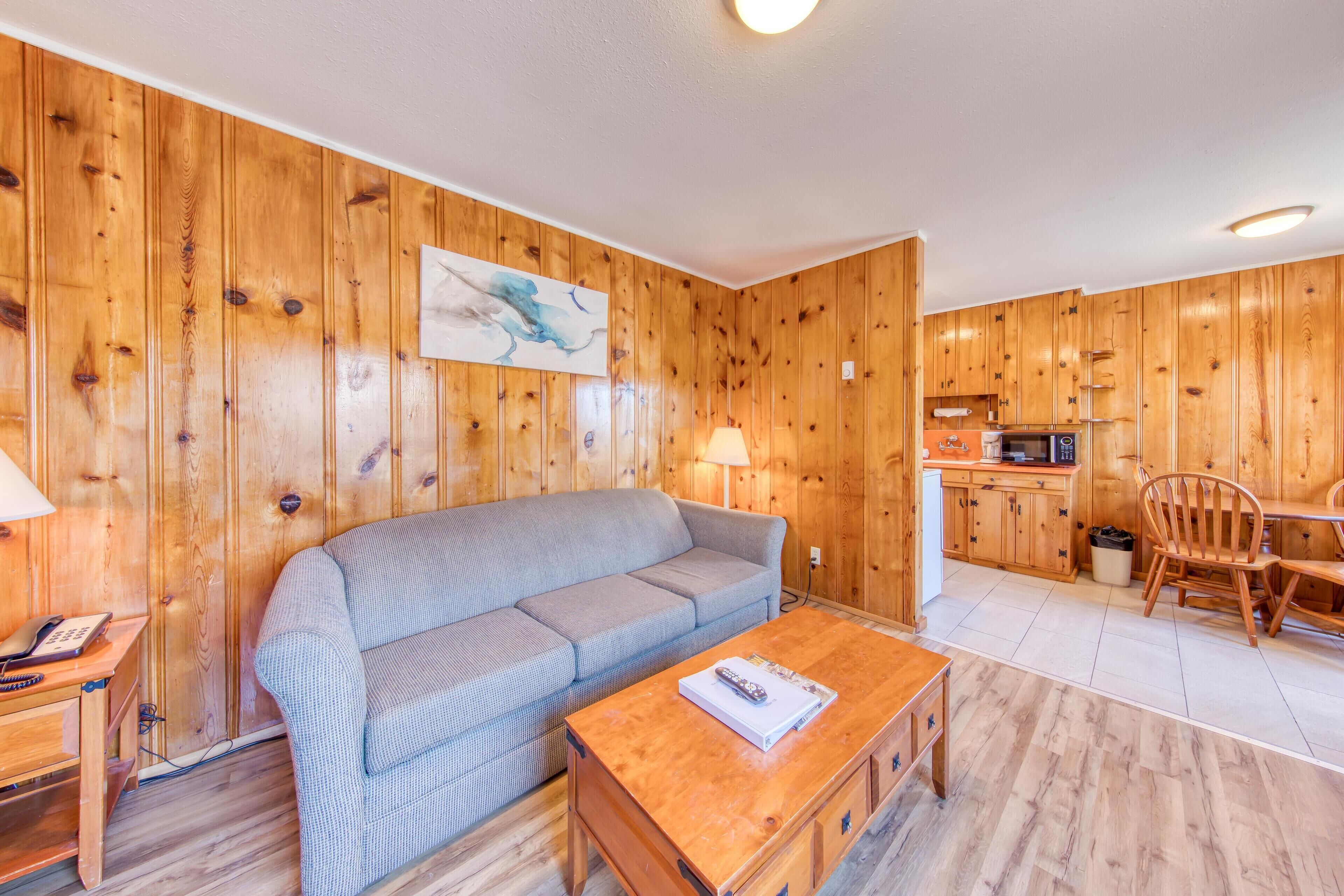Una habitación acogedora con una pared de madera, un sofá gris, una mesa de centro de madera y una zona de cocina con armarios de madera y un microondas.