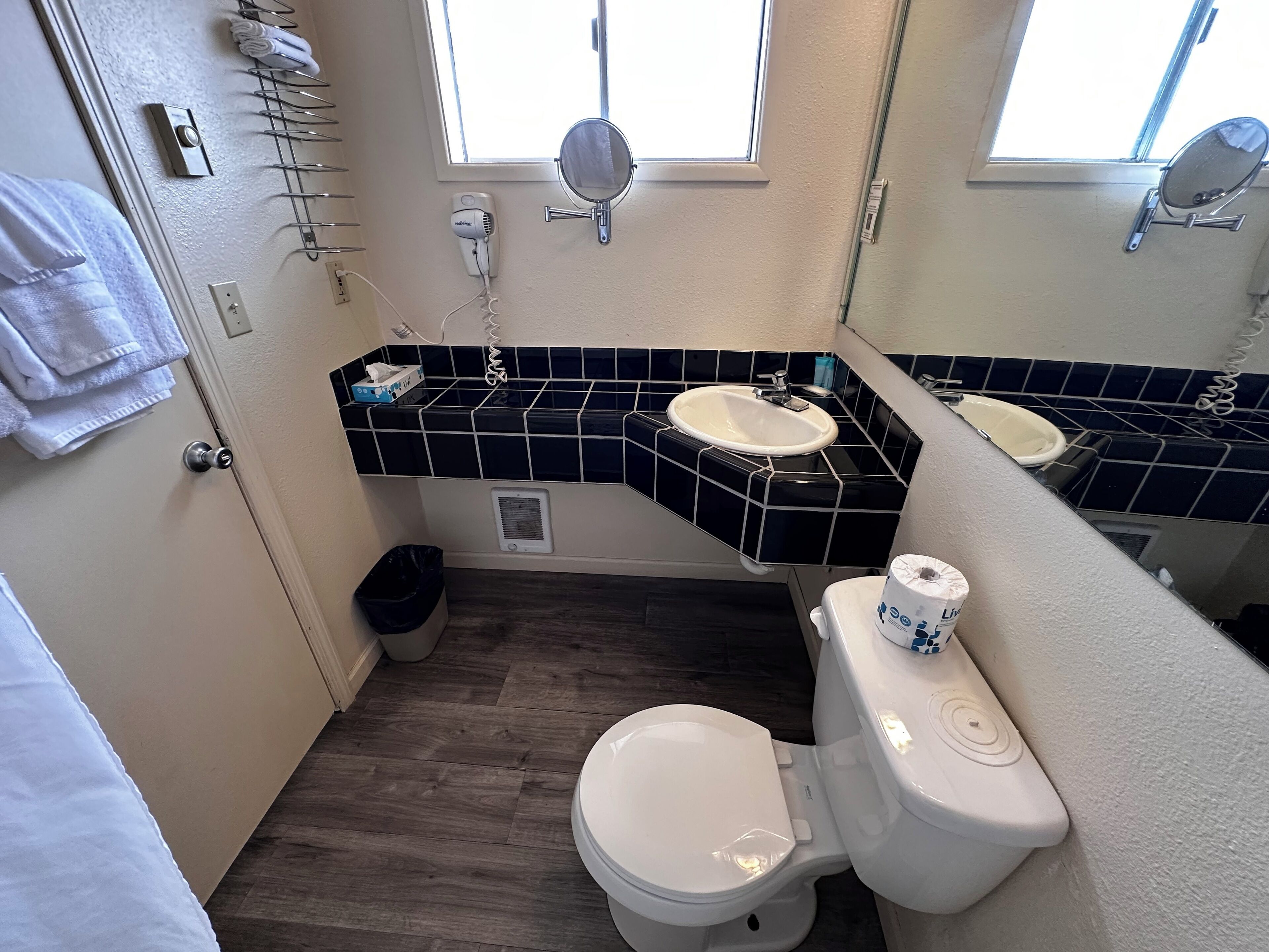 A bathroom with a white toilet, a sink, and a mirror. There are towels hanging on the wall, a trash can, and a window providing natural light.