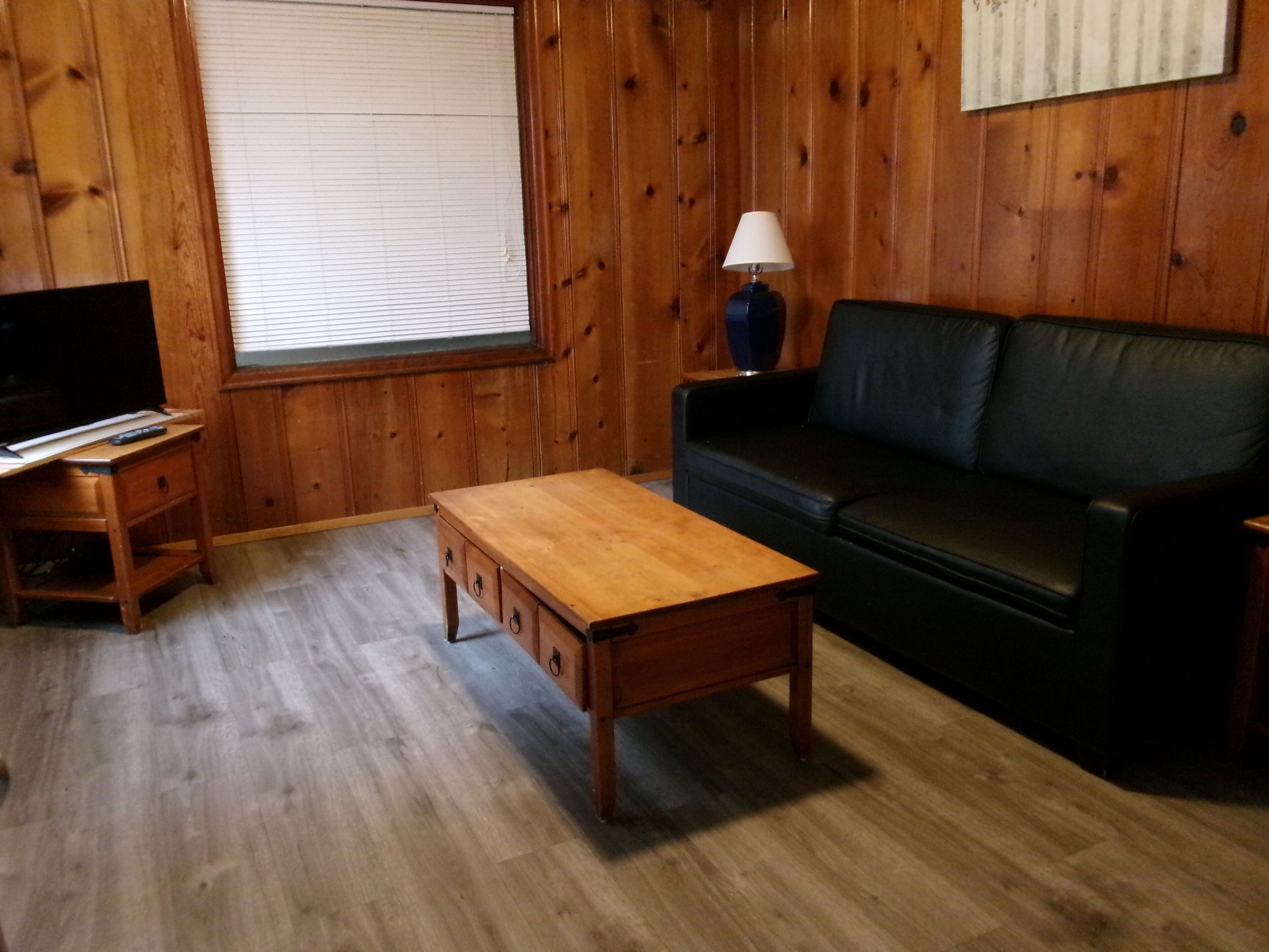 A wooden-paneled room with a black leather sofa, a wooden coffee table, a TV stand, and a lamp.