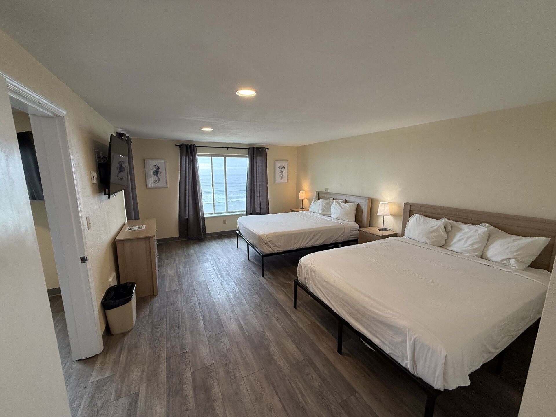 A hotel room with two beds, a TV, a window with curtains, and a view of the sea.