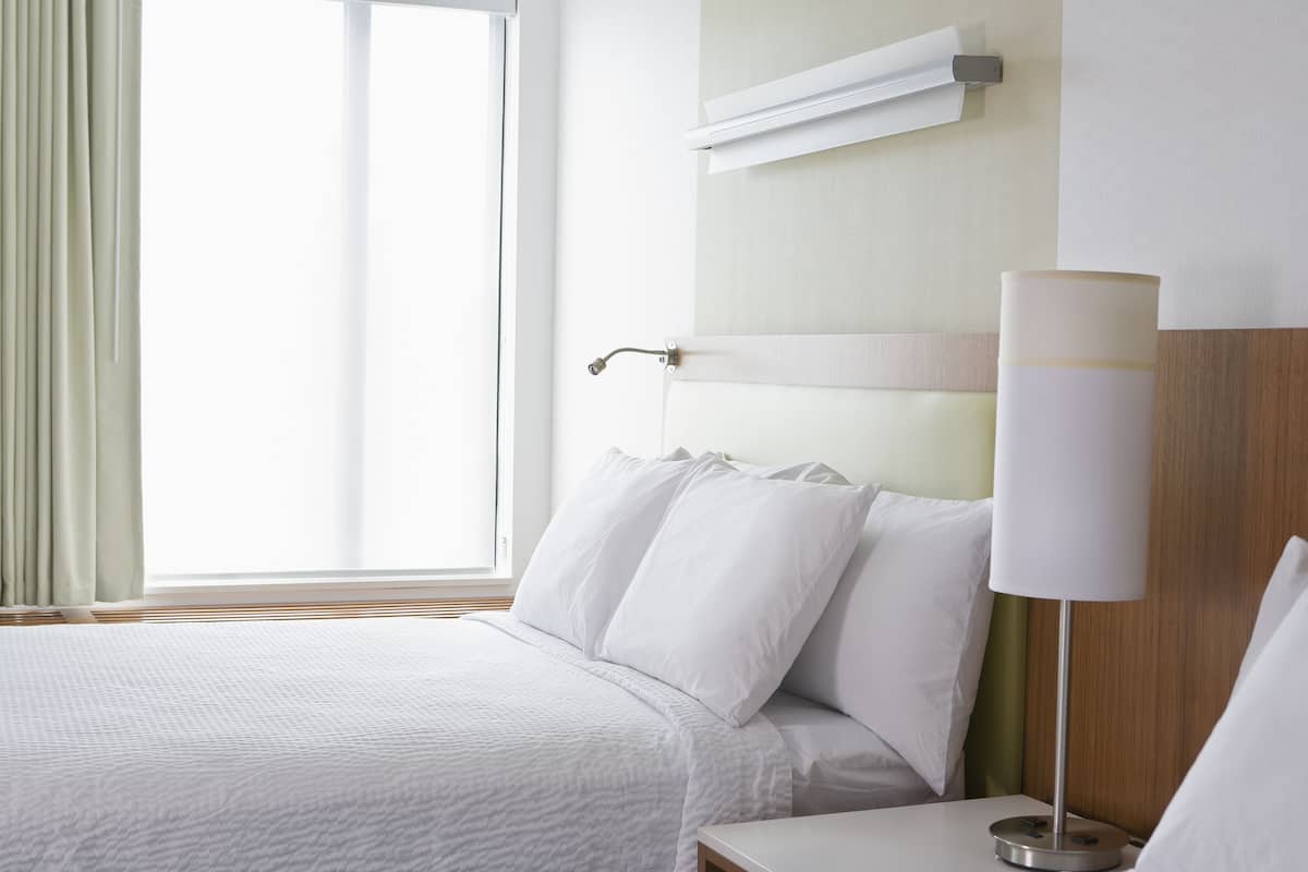 Una habitación de hotel con cama, mesita de noche, lámpara y ventana con cortinas.