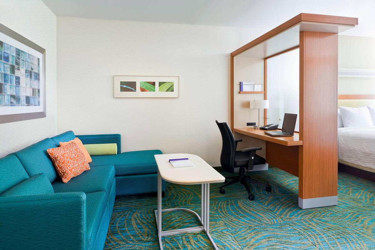 A compact hotel room with a teal sofa, a small desk with a laptop, a chair, and a bed.