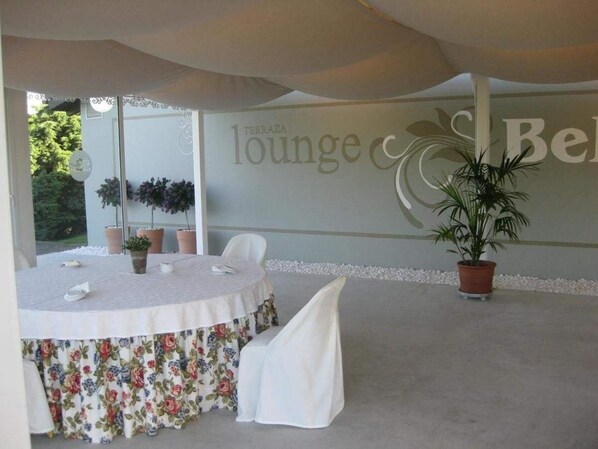 Una mesa redonda con mantel floral y sillas blancas, una planta en maceta y una pared con el texto "Terraza lounge cBe".