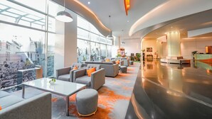 Un hall d’entrée spacieux au design moderne, comprenant un comptoir d’accueil, un espace salon avec des canapés et des fauteuils, et de grandes fenêtres offrant une vue sur l’extérieur.