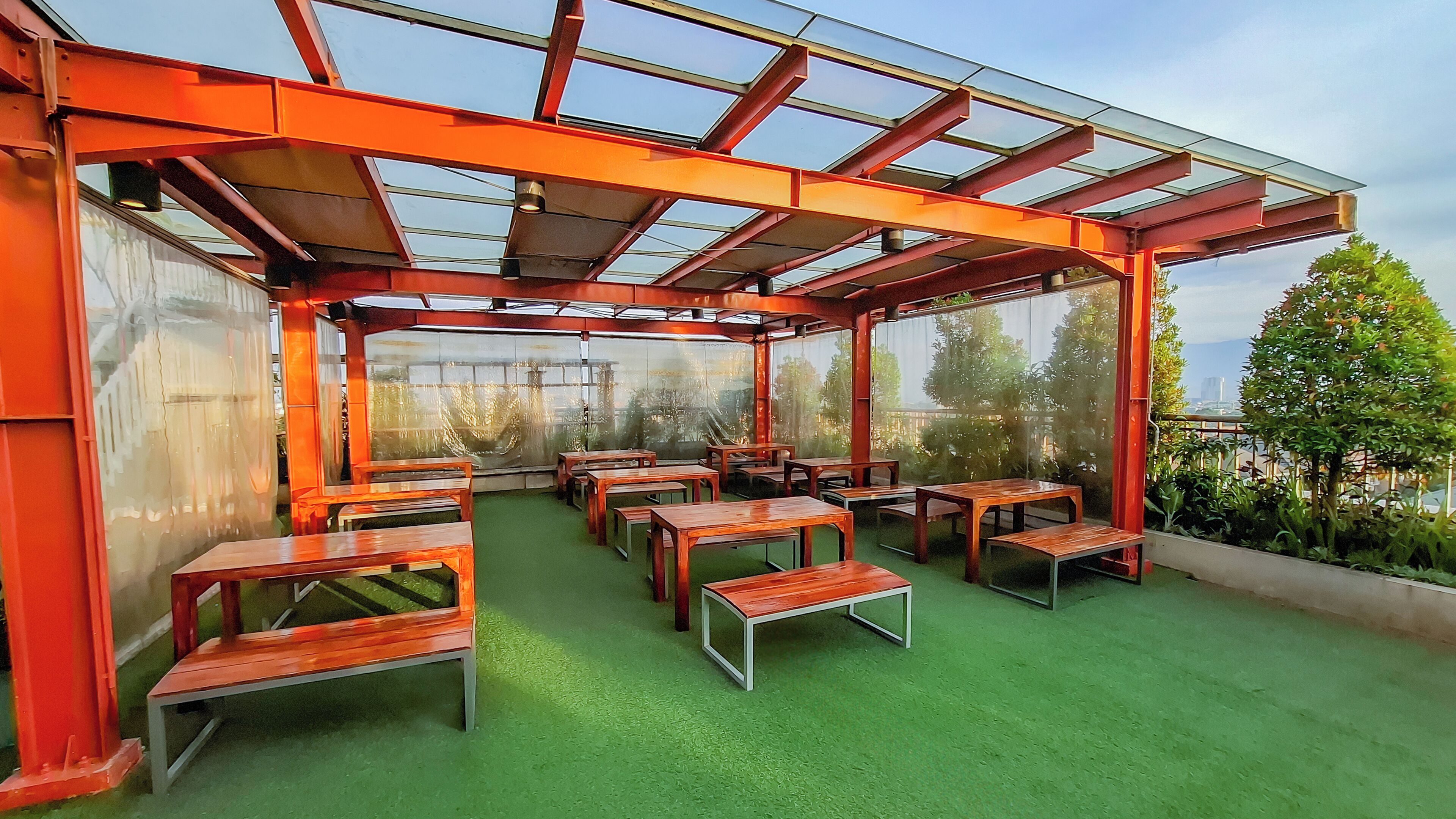 A covered outdoor seating area with tables and benches, surrounded by glass walls and a green floor.