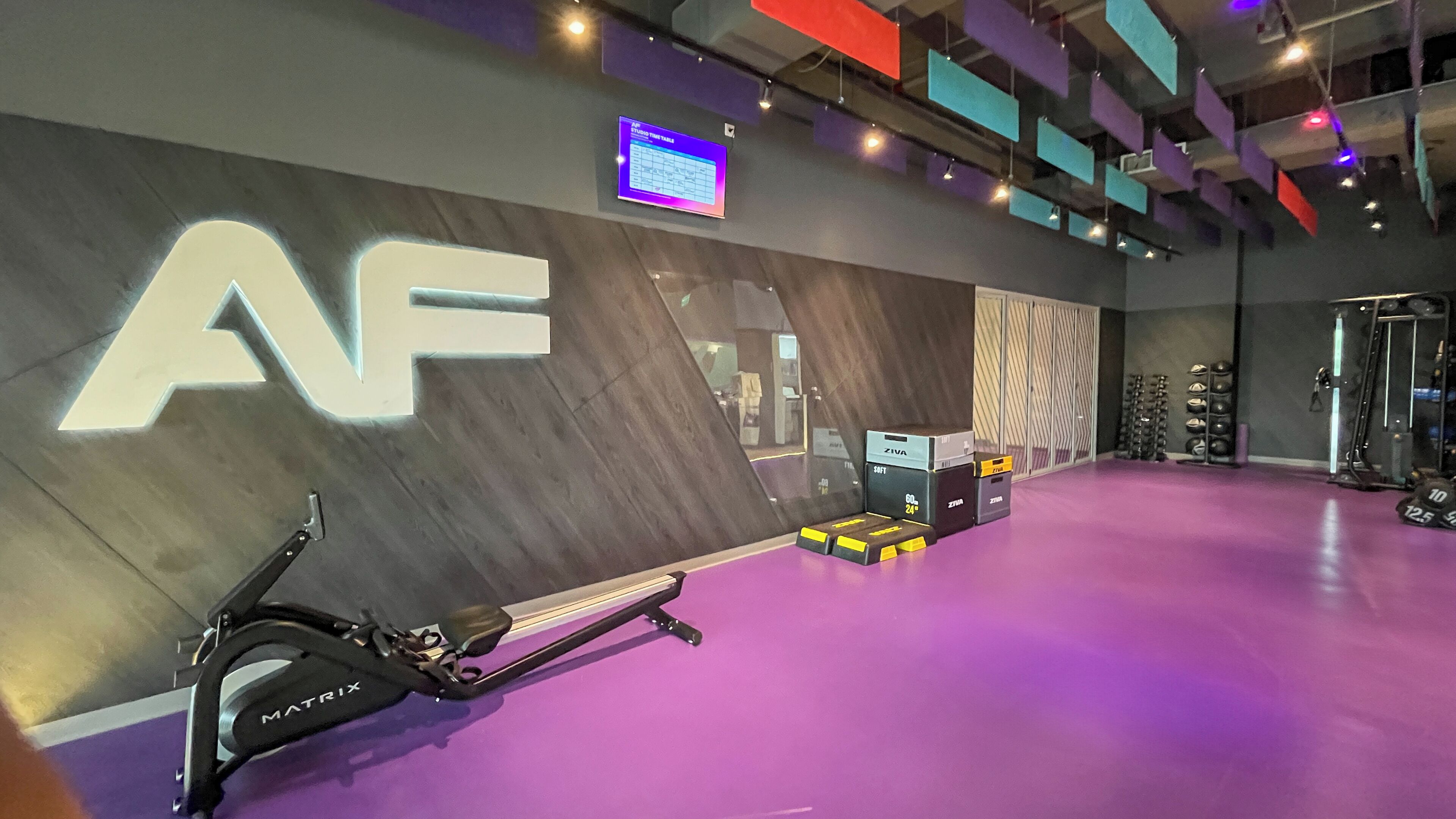 Uma academia com um grande letreiro 'AF', equipamentos de exercícios e uma tela digital na parede.