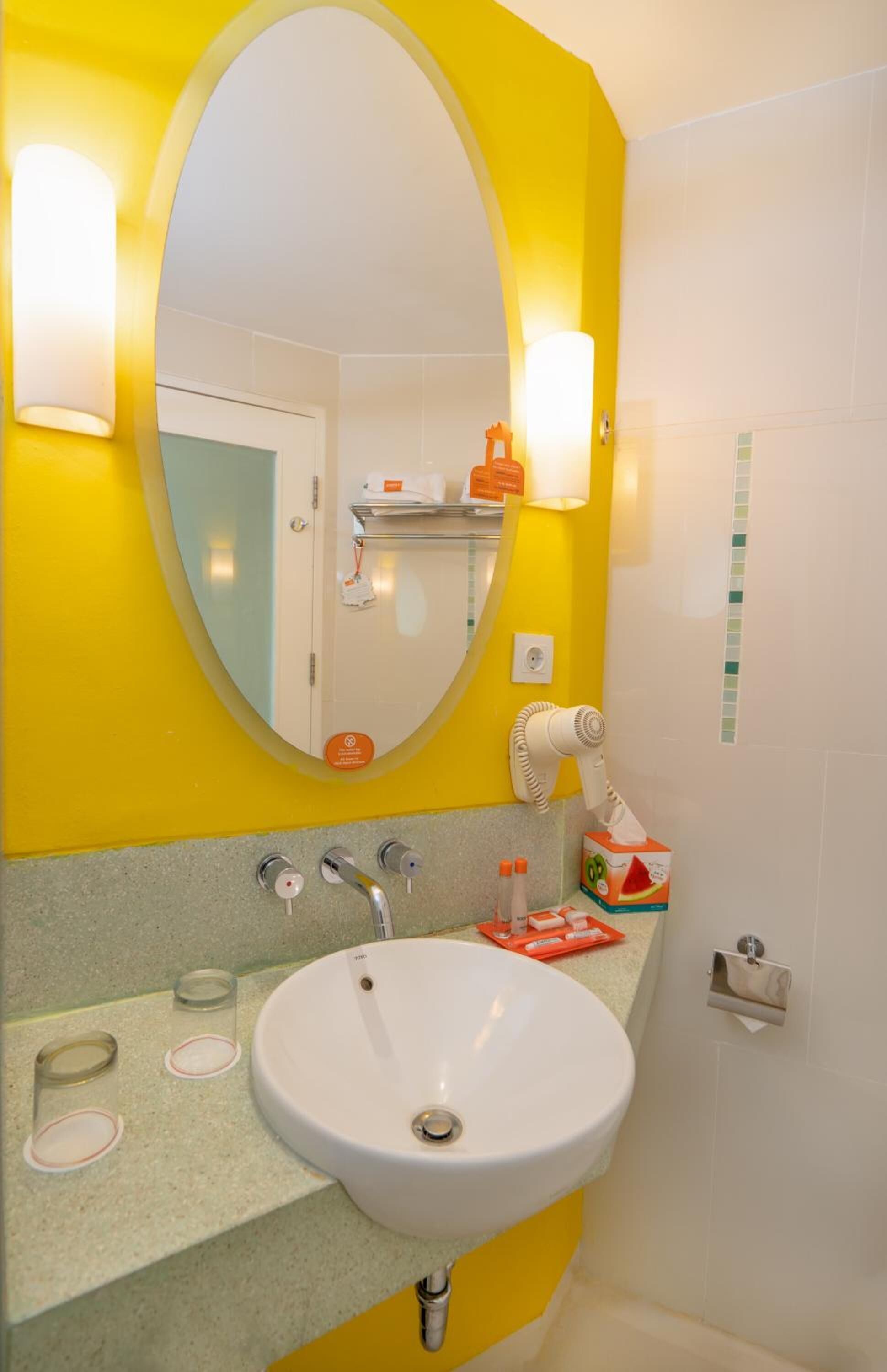 Une salle de bain aux couleurs jaune et blanc, avec un grand miroir ovale, un lavabo blanc et un porte-serviettes.