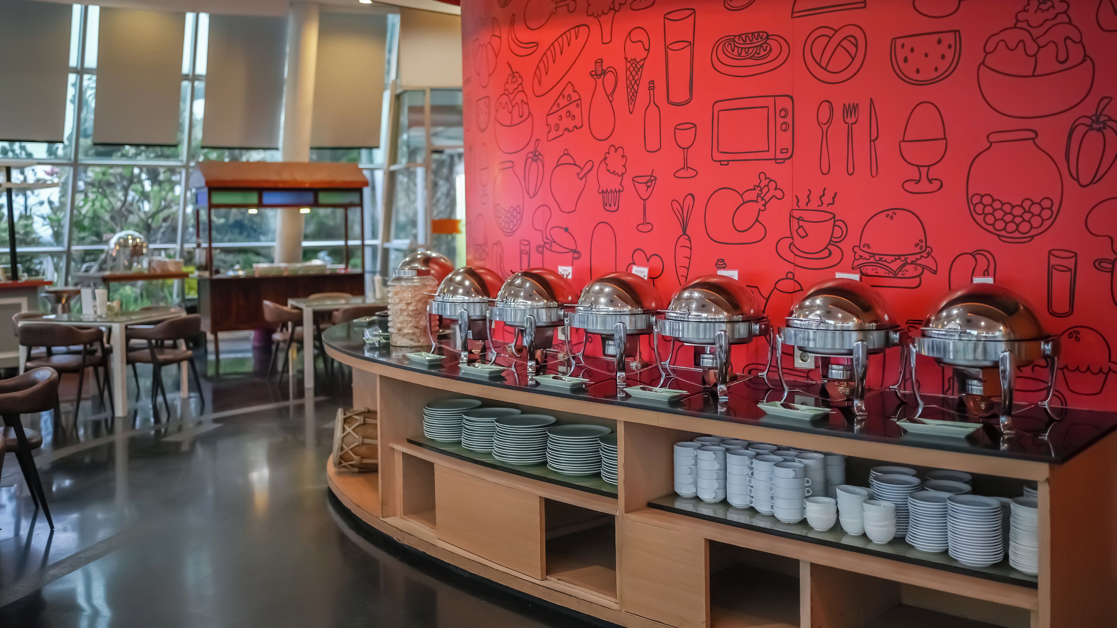 Zona de bufé con varios dispensadores de comida, una barra con platos apilados y una pared roja decorada con ilustraciones de alimentos.