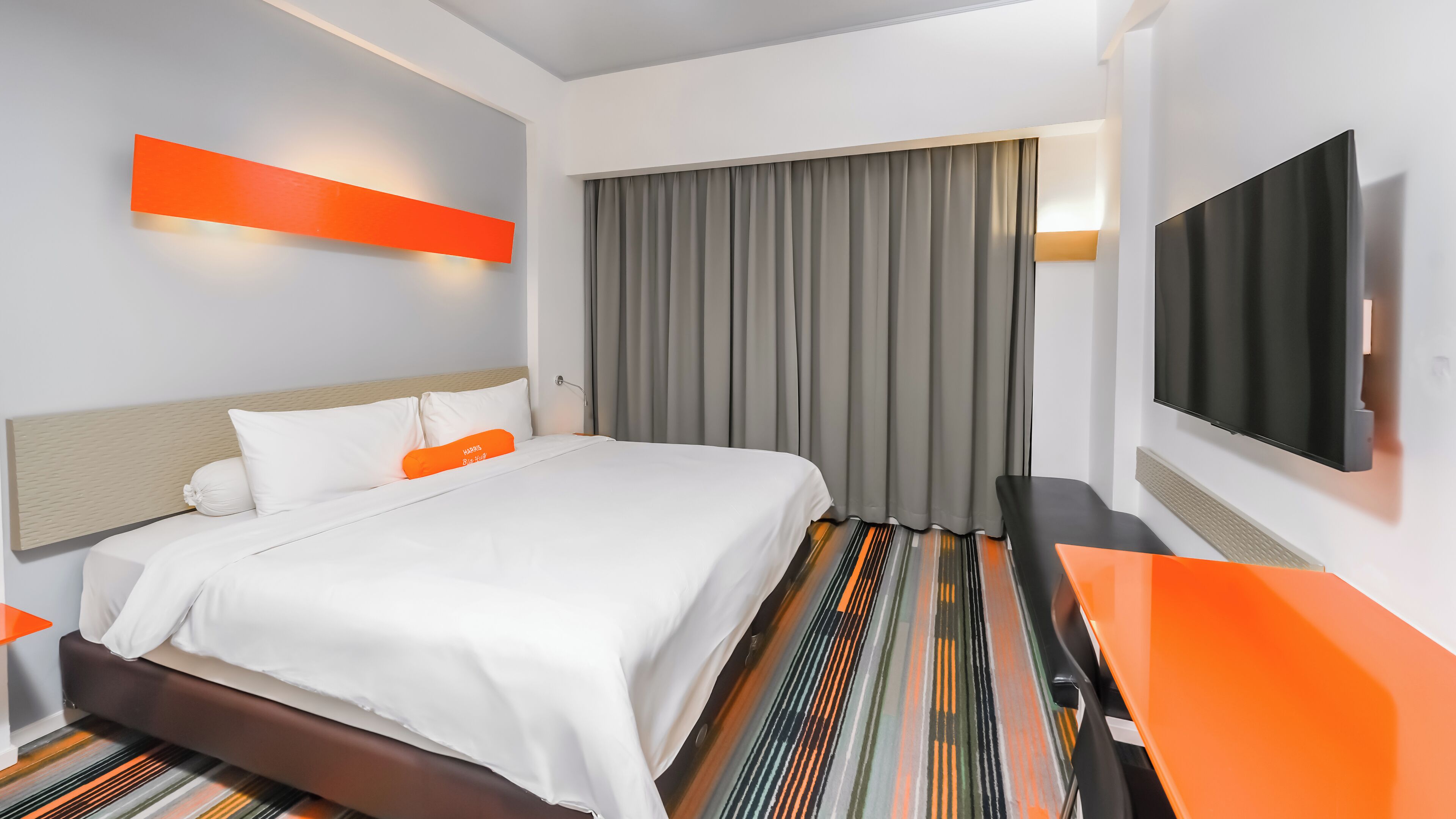 Hotellihuone, jossa on sänky, televisio, työpöytä ja suuri ikkuna, jossa on verhot.