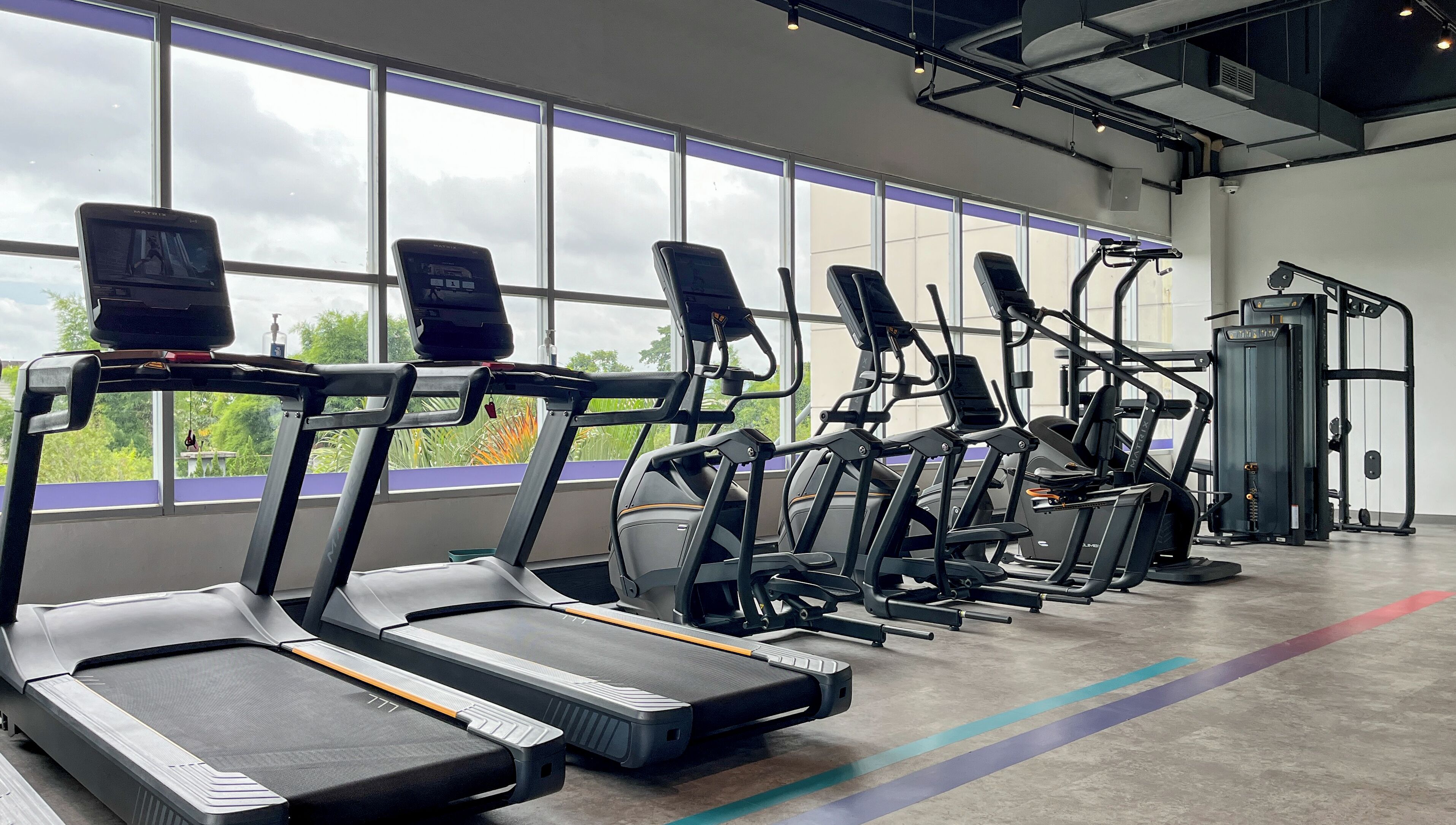 A gym with treadmills and exercise equipment, large windows, and a concrete floor.
