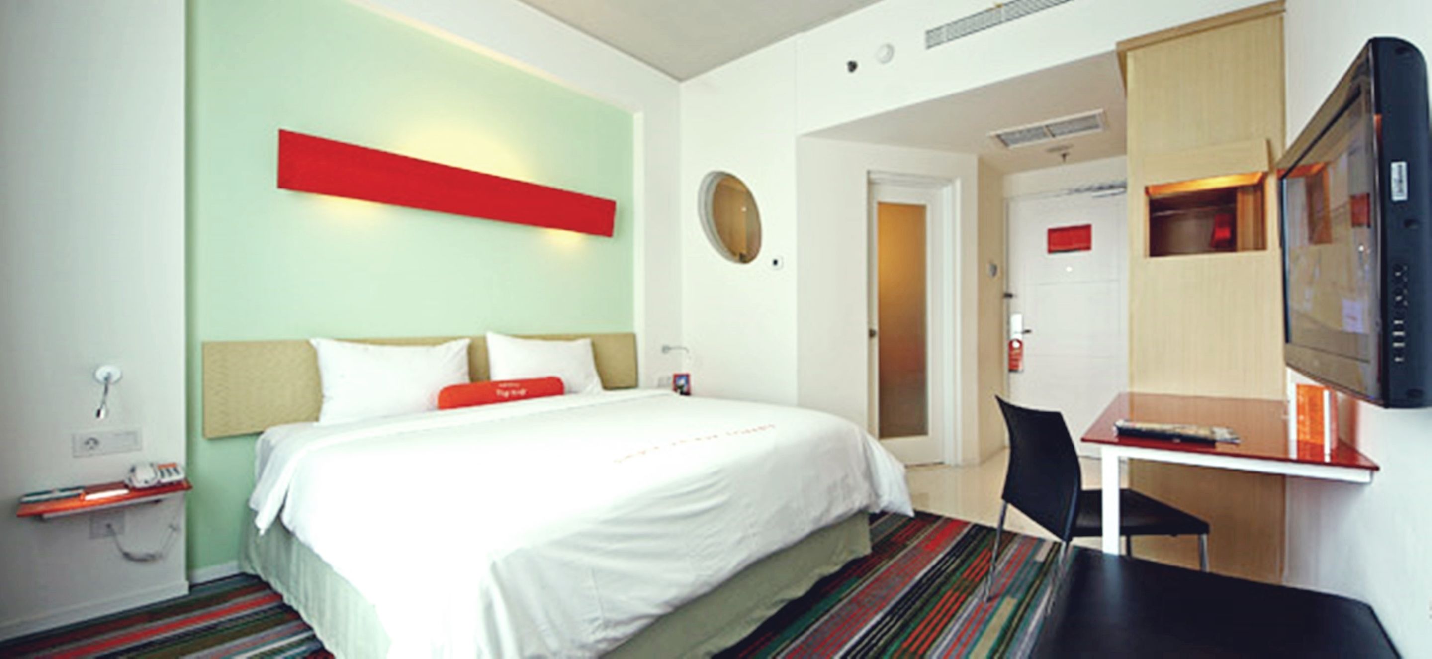 A hotel room with a bed, a desk, a chair, a TV, and a door leading to another room.