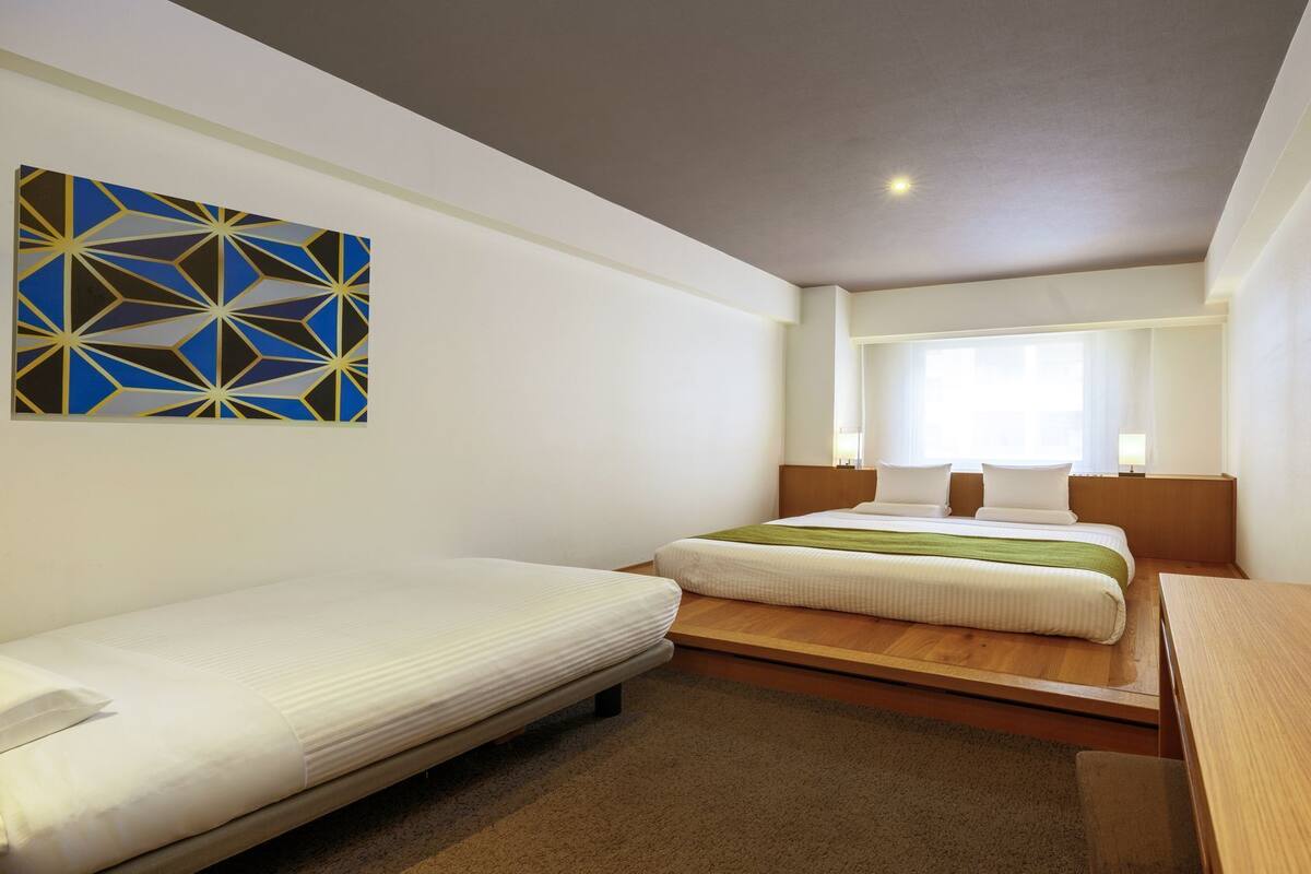 A hotel room with two beds, a large window, a flat-screen TV, and a geometric artwork on the wall.