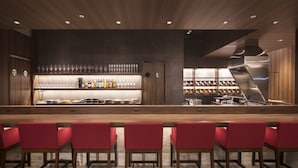 A bar area with a long counter, red bar stools, and shelves with bottles and glasses.