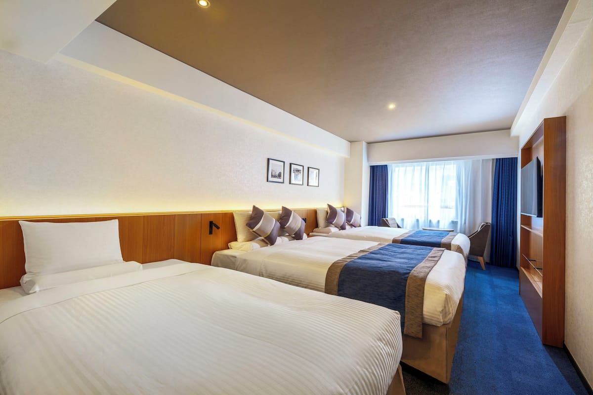 A hotel room with three beds, a wooden headboard, a flat-screen TV, and a large window with curtains.