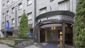 A hotel entrance with a curved facade, a prominent sign reading "HOTEL MYSTAYS PREMIER", and a blue carpet leading to the entrance.