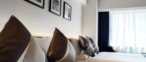 A hotel room with a neatly arranged bed, multiple pillows, a large window with curtains, and framed pictures on the wall.