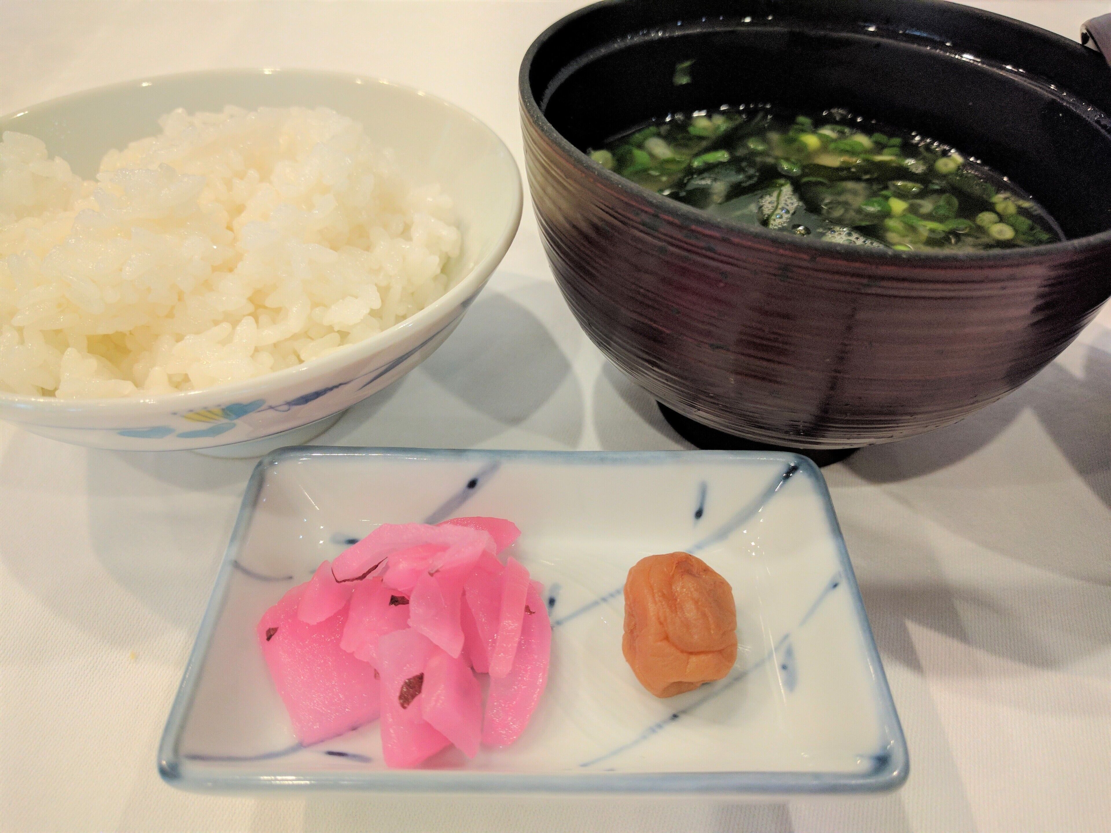 A bowl of white rice, a bowl of green vegetable soup, and a small dish with pink pickled vegetables and a piece of orange food on a table.