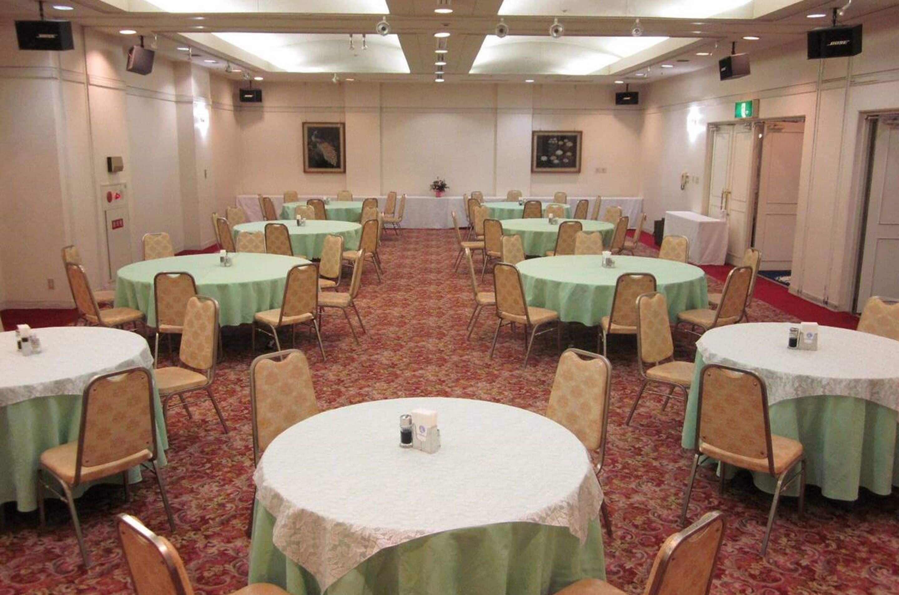A conference room with multiple round tables covered with green tablecloths, beige chairs, and a carpeted floor.