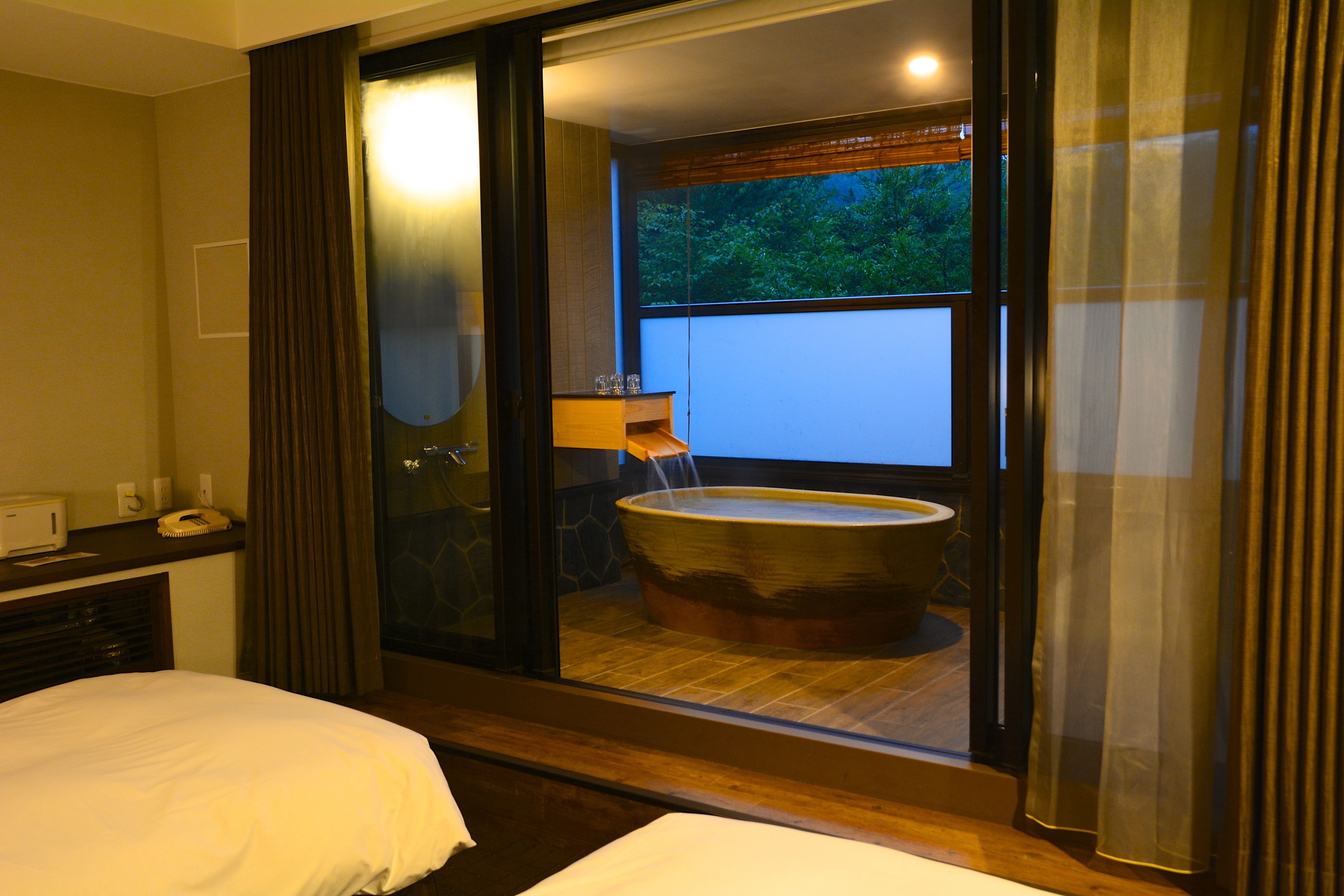 A hotel room with a large bathtub, a bed, a desk with a telephone, and a window with a view of greenery.