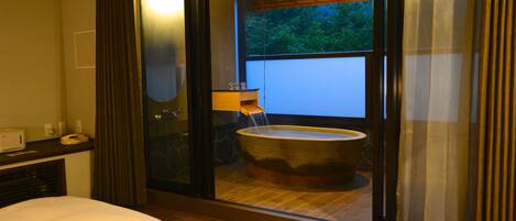 A hotel room with a large bathtub, a bed, a desk with a telephone, and a window with a view of greenery.