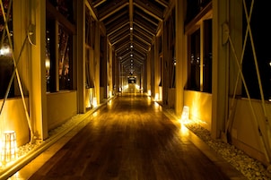A corridor with wooden flooring, illuminated by wall-mounted lights, flanked by windows and white railings.