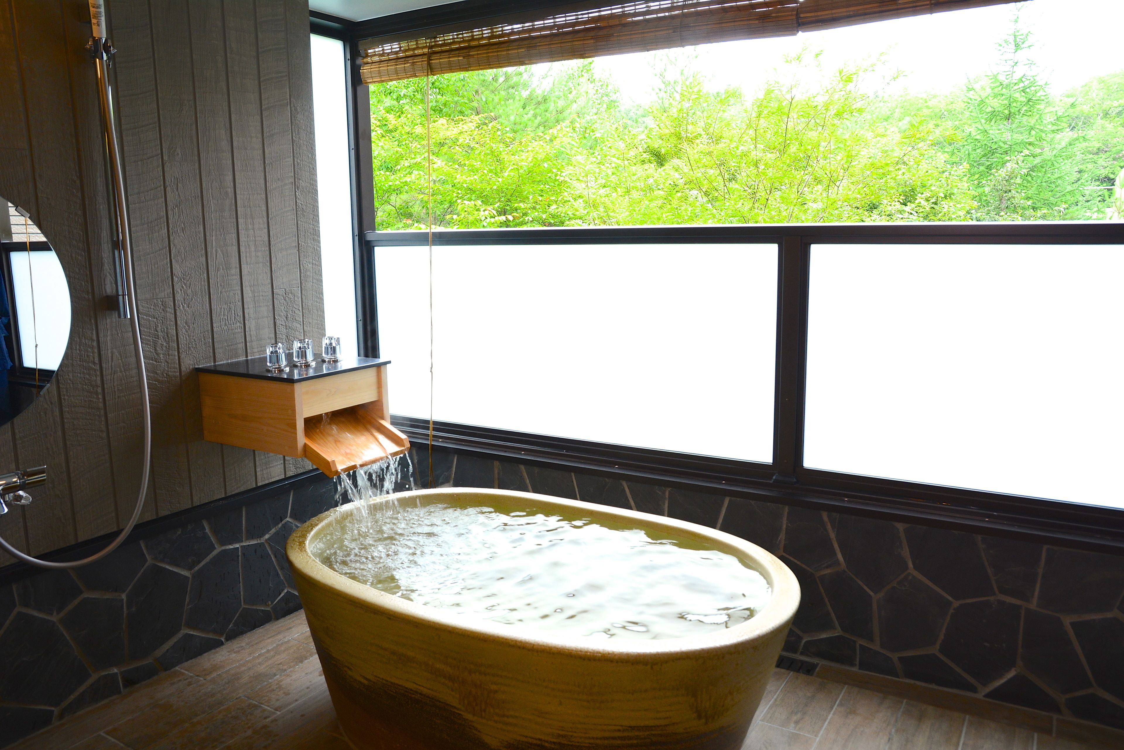 A bathtub with water flowing into it, a wooden shelf with glasses, and a window with a view of greenery.