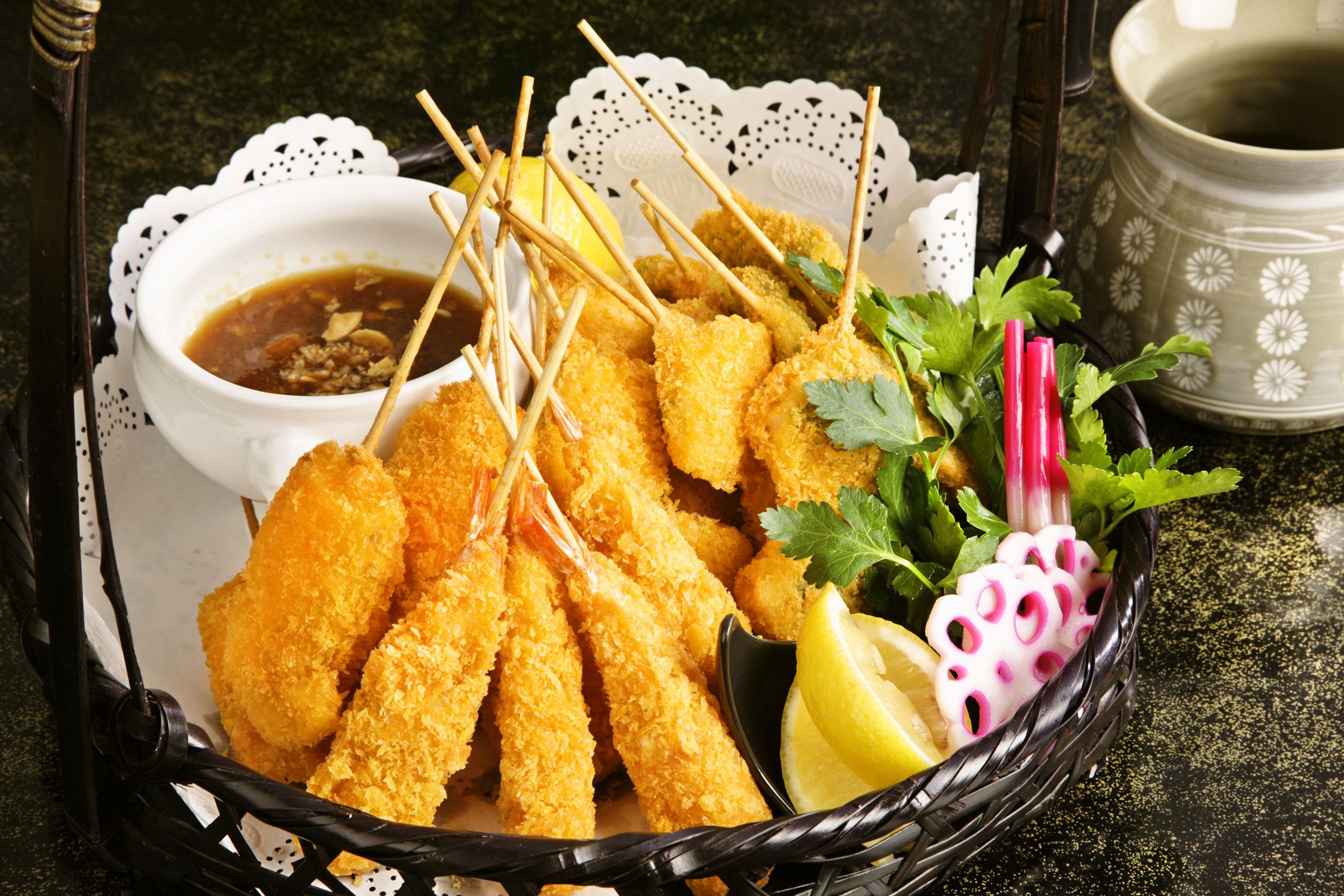 Um cesto com espetinhos de camarão frito, fatias de limão e uma pequena tigela de molho.